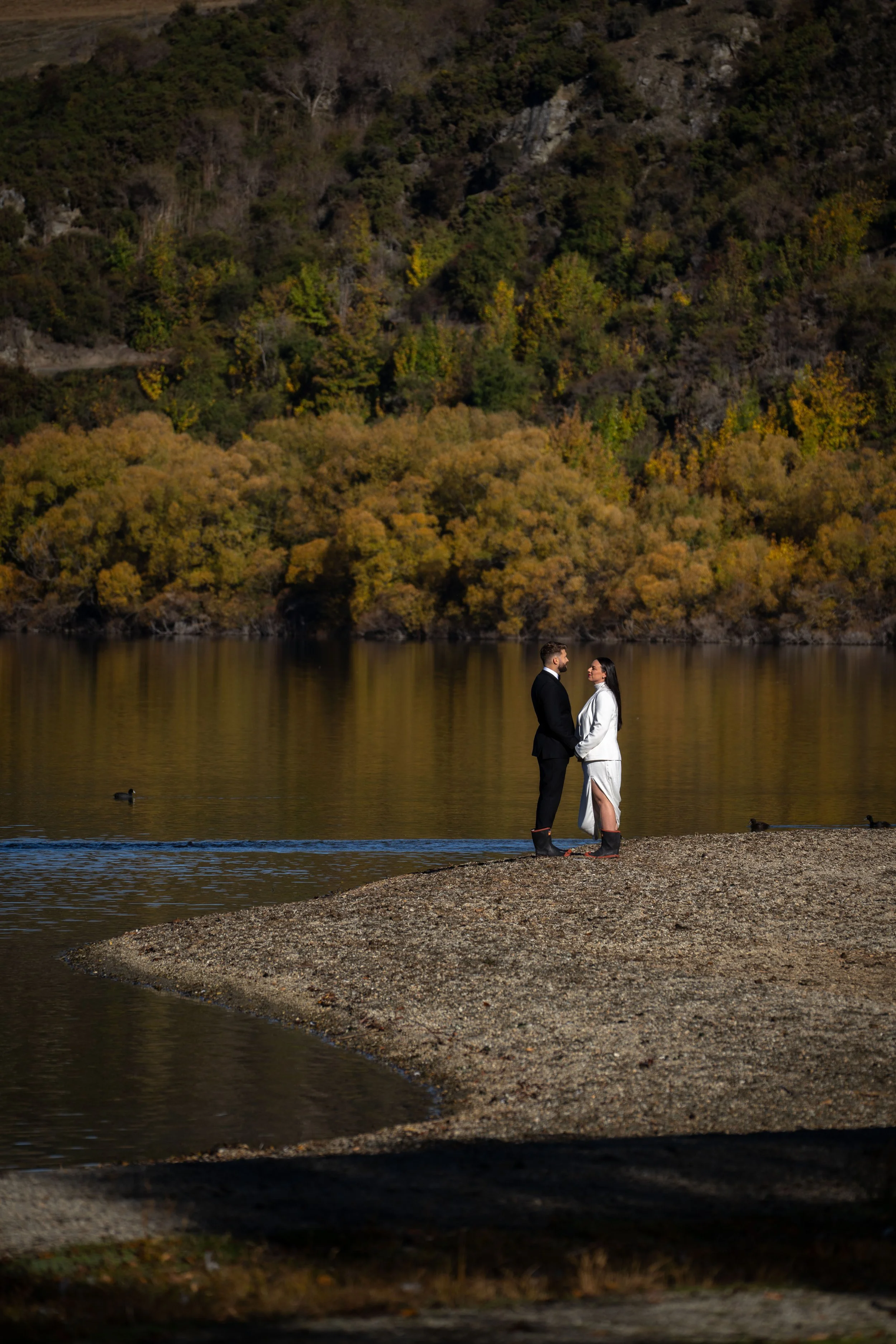 A couple dressed in formal wedding attire holding hands and standing facing each other on a narrow strip of rocky land by a lake, with a backdrop of colorful autumn trees on a hillside.
