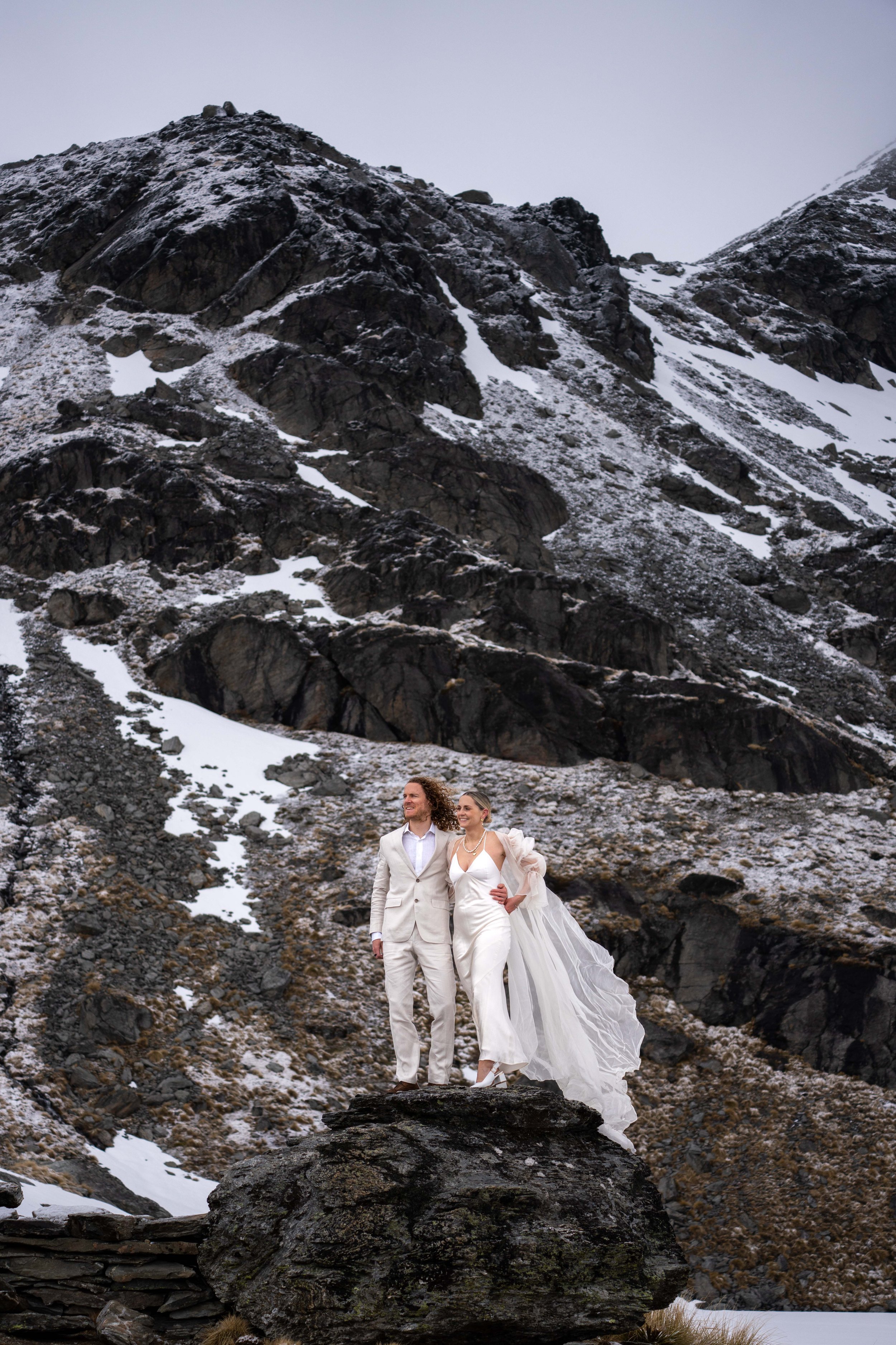 A newlywed couple stands on a large rock in a snowy mountainous landscape. The bride wears a white wedding dress with a long flowing train, and the groom is dressed in a light-colored suit. They are smiling and holding each other, enjoying the scenic
