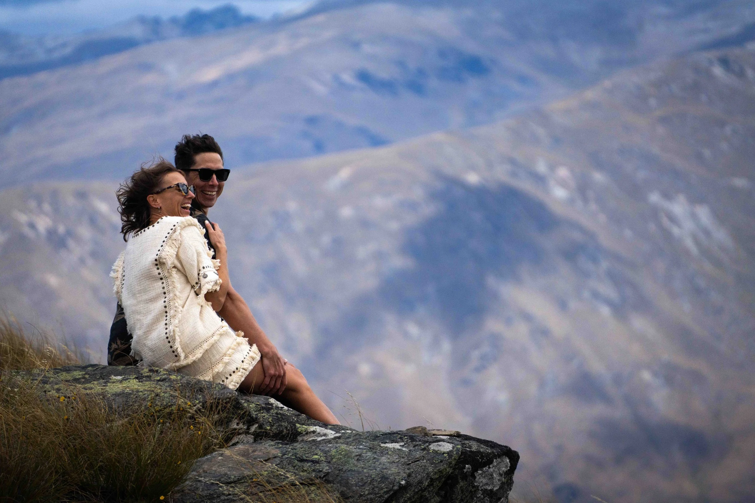 A couple sitting on a rock, smiling and laughing with mountains in the background.