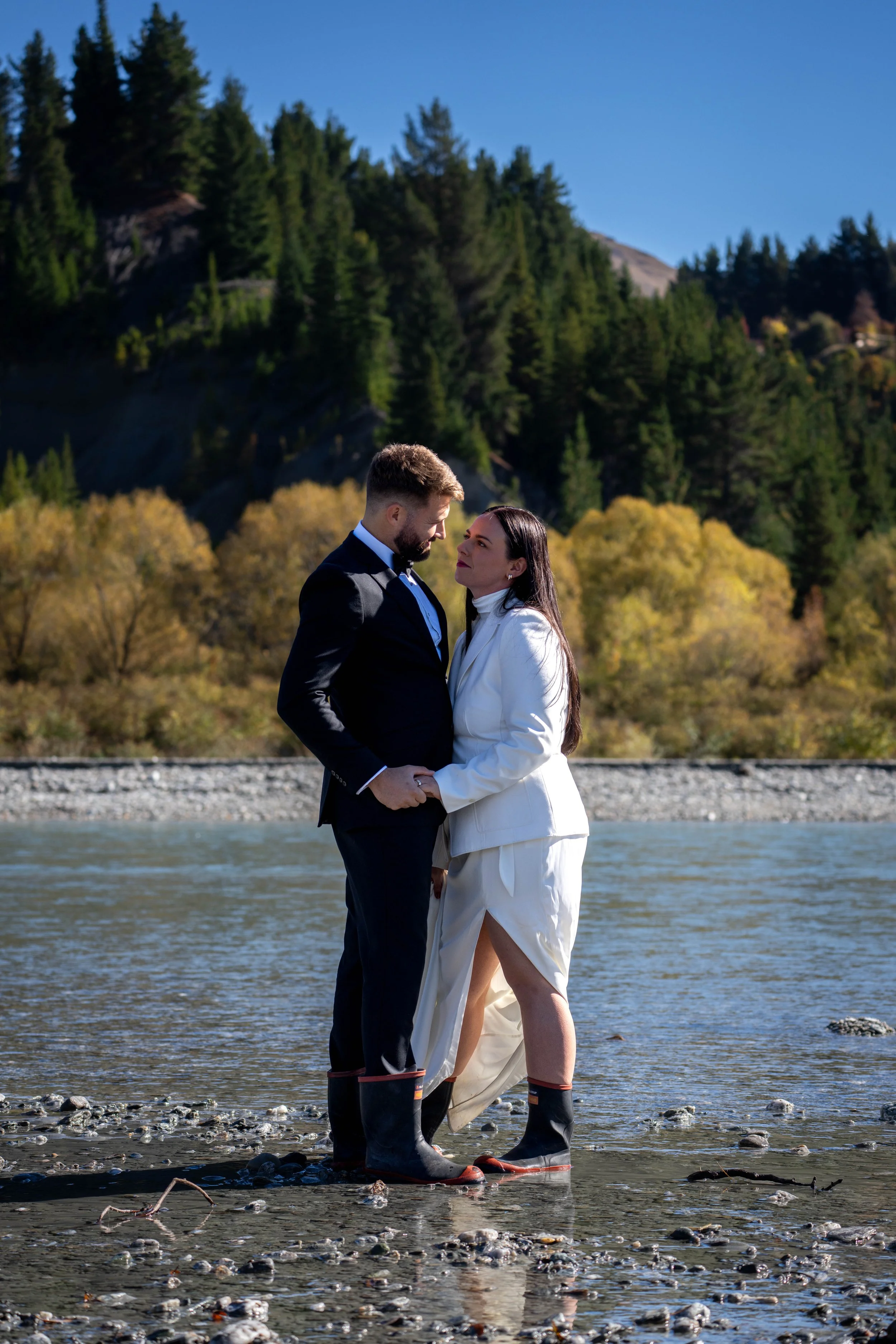 A couple dressed in formal attire stands in a shallow river, holding hands and looking at each other with a backdrop of trees and mountains.