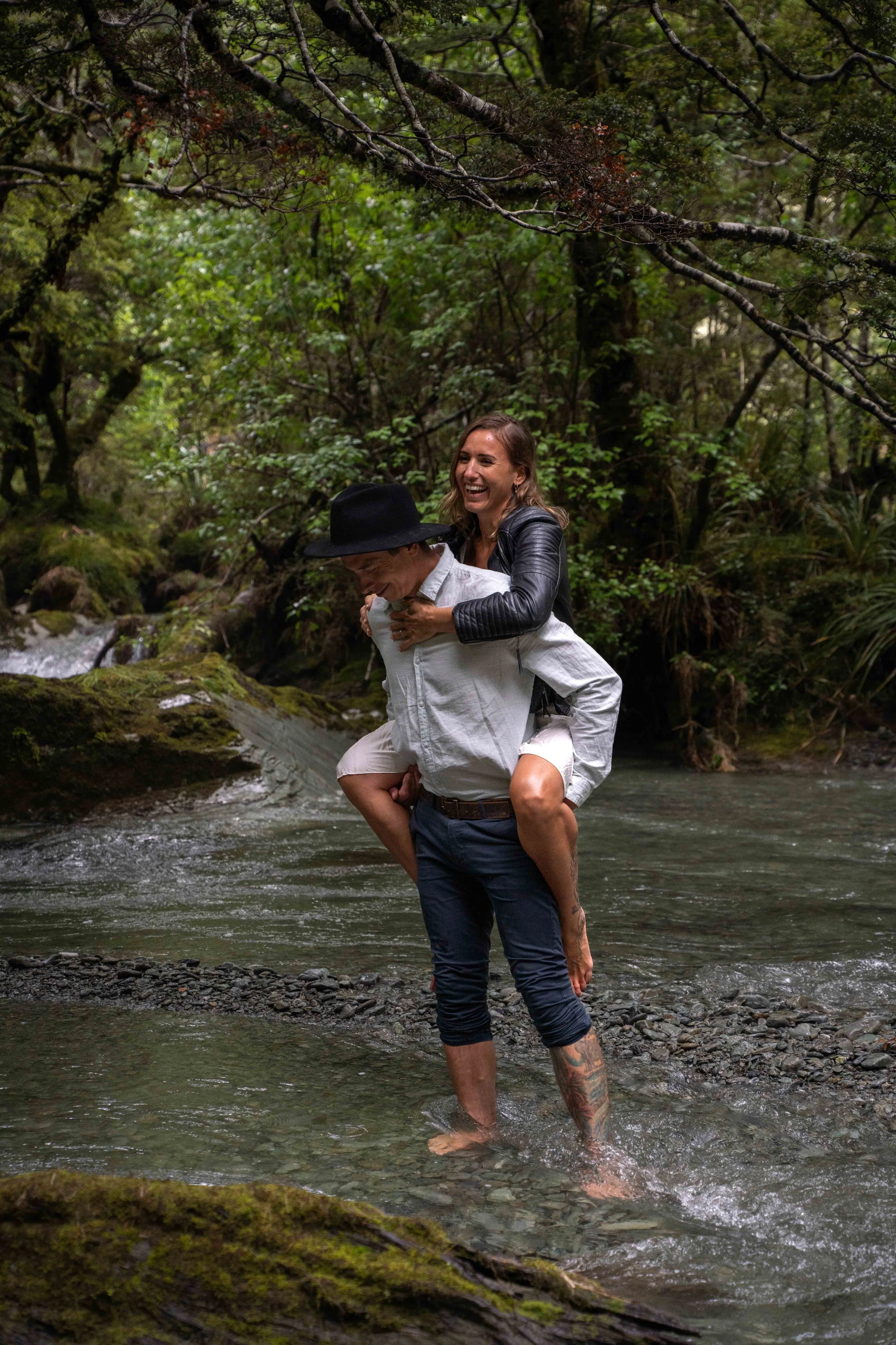 A man giving a woman a piggyback ride in a forest stream, both are smiling and enjoying nature.