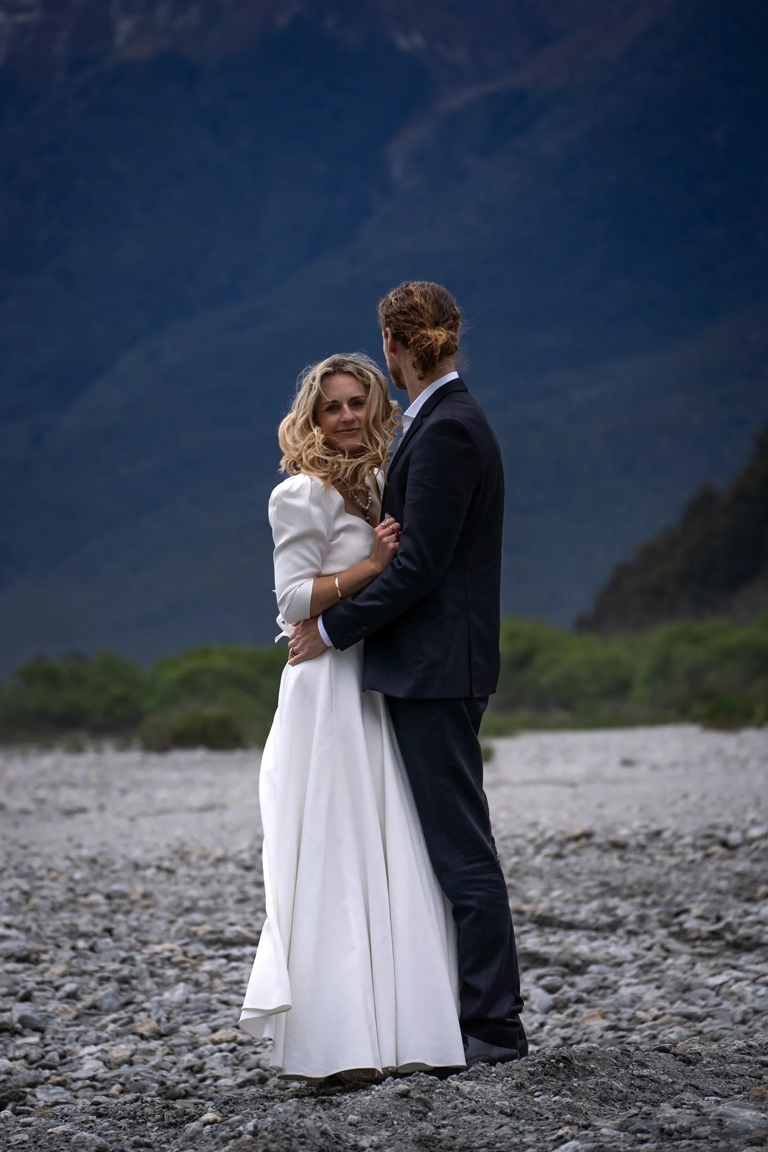 A couple dressed in formal wedding attire embracing outdoors on a rocky terrain with mountains and a dark sky in the background.