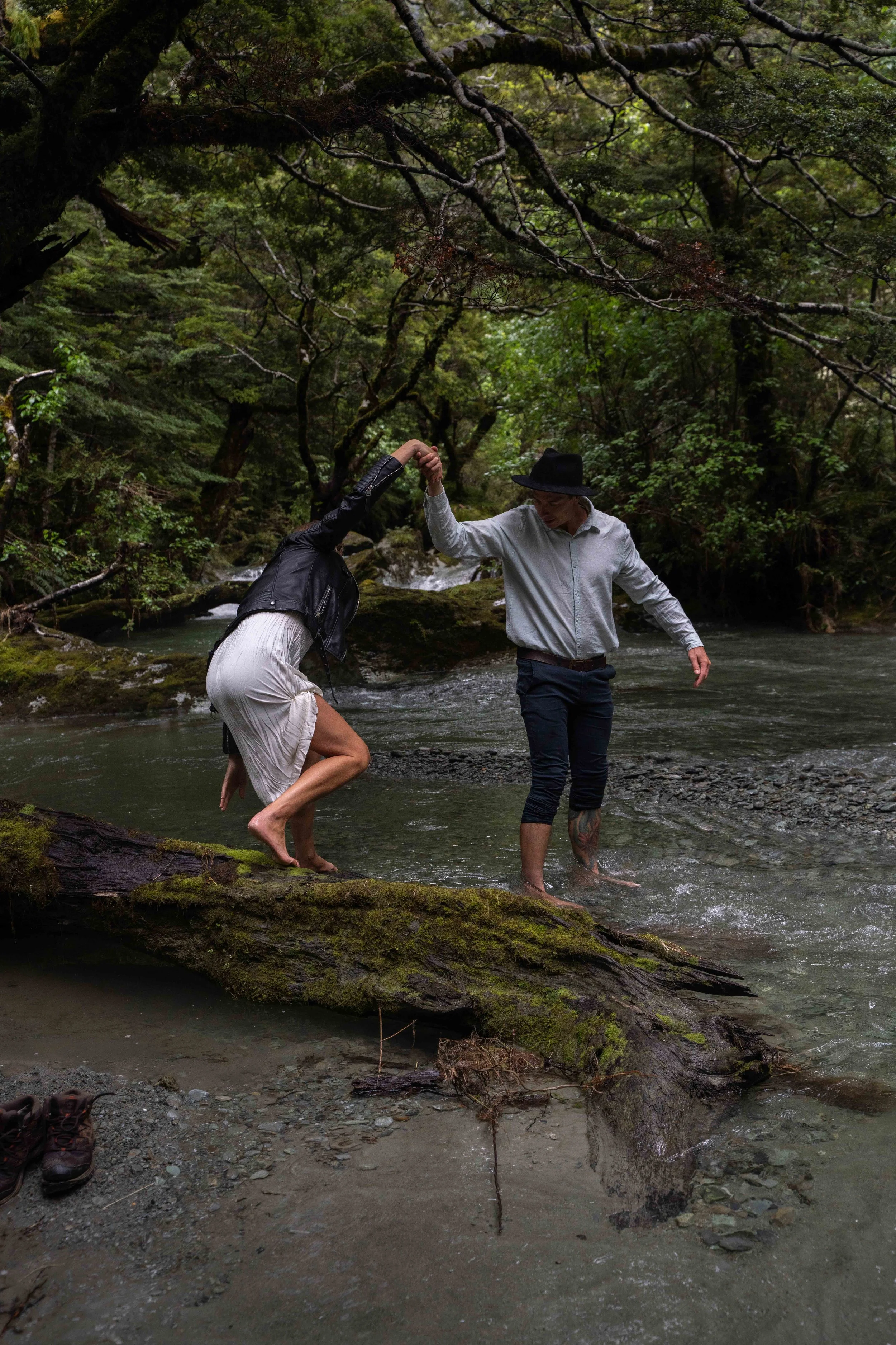 A couple climbing over a fallen log in a forest stream, with lush green trees surrounding them.