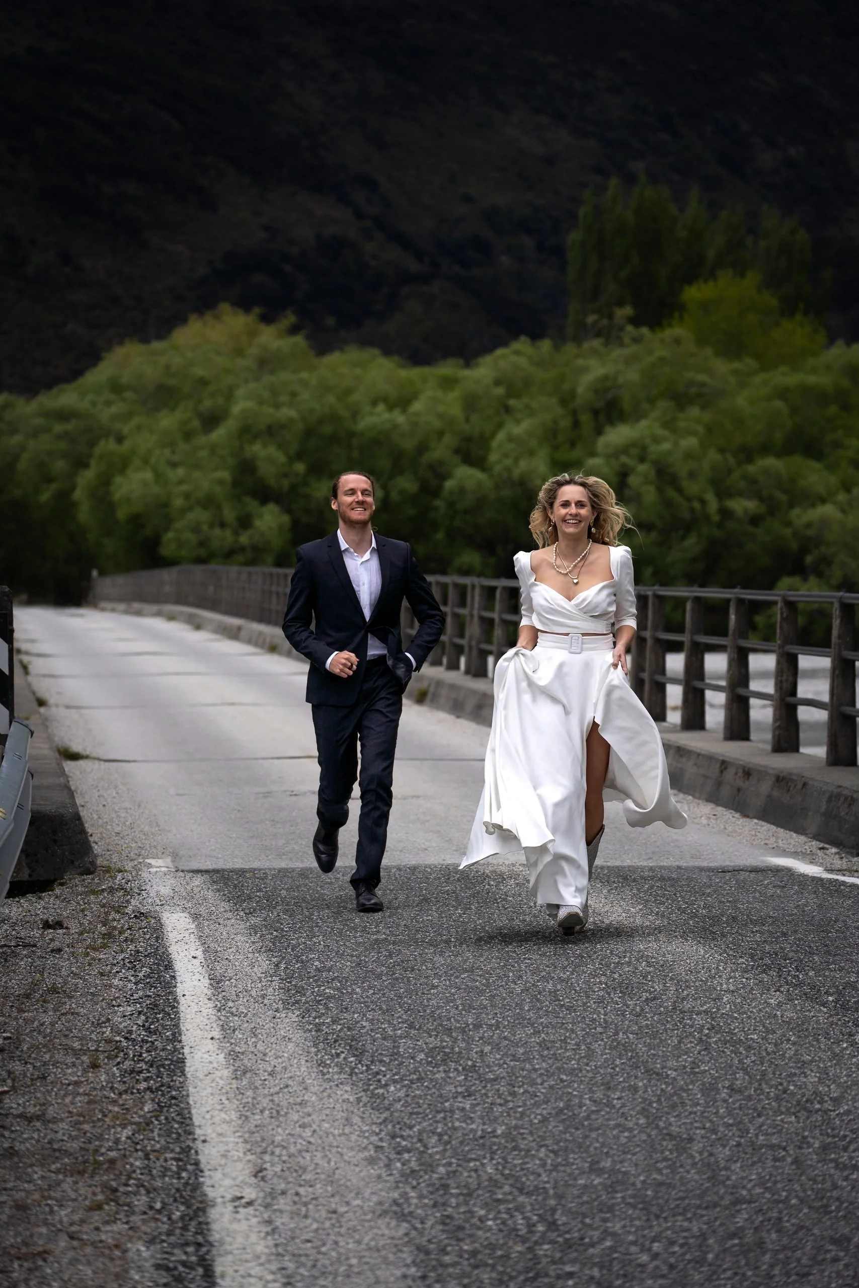 A bride and groom running on a bridge in a scenic outdoor setting, with lush green trees and mountains in the background.
