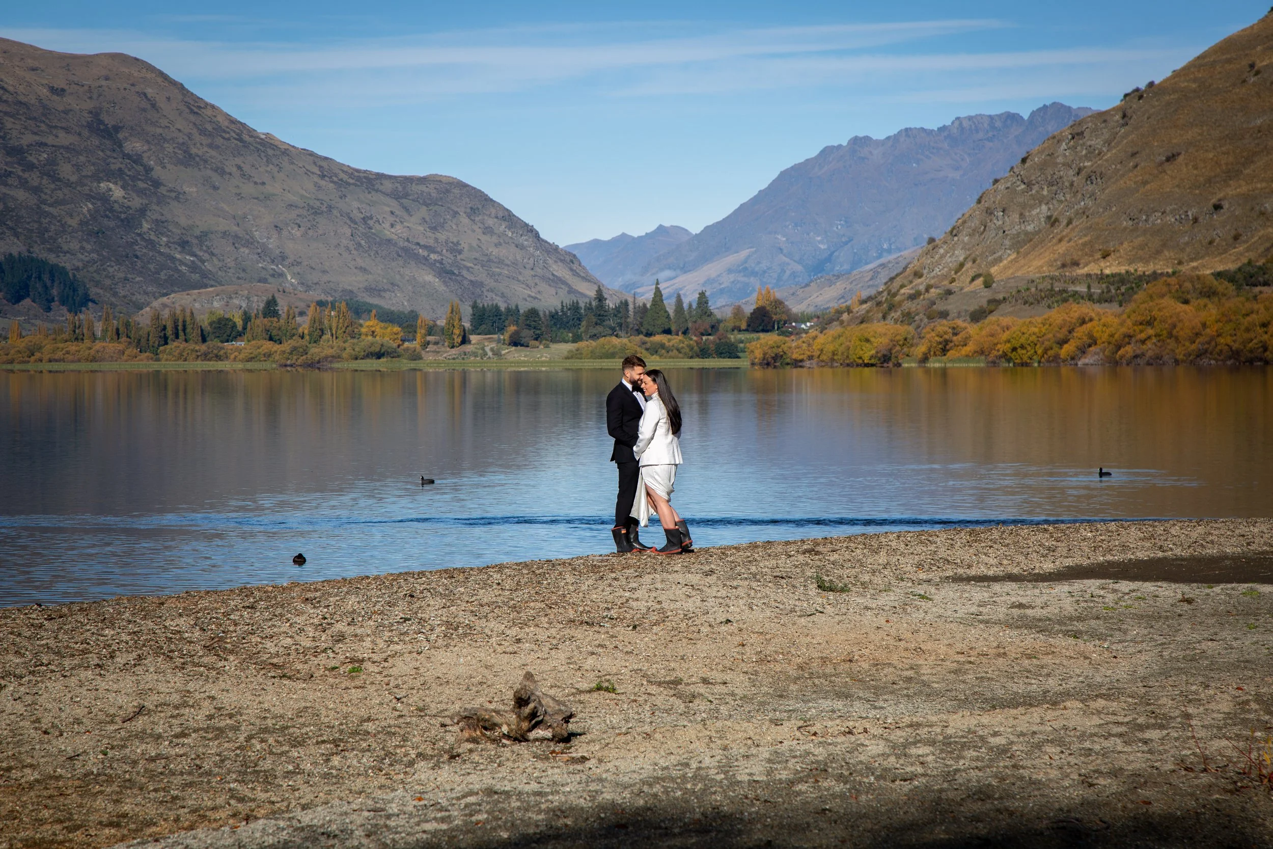 A couple dressed in formal attire embracing by a lake in a mountainous landscape during daytime.