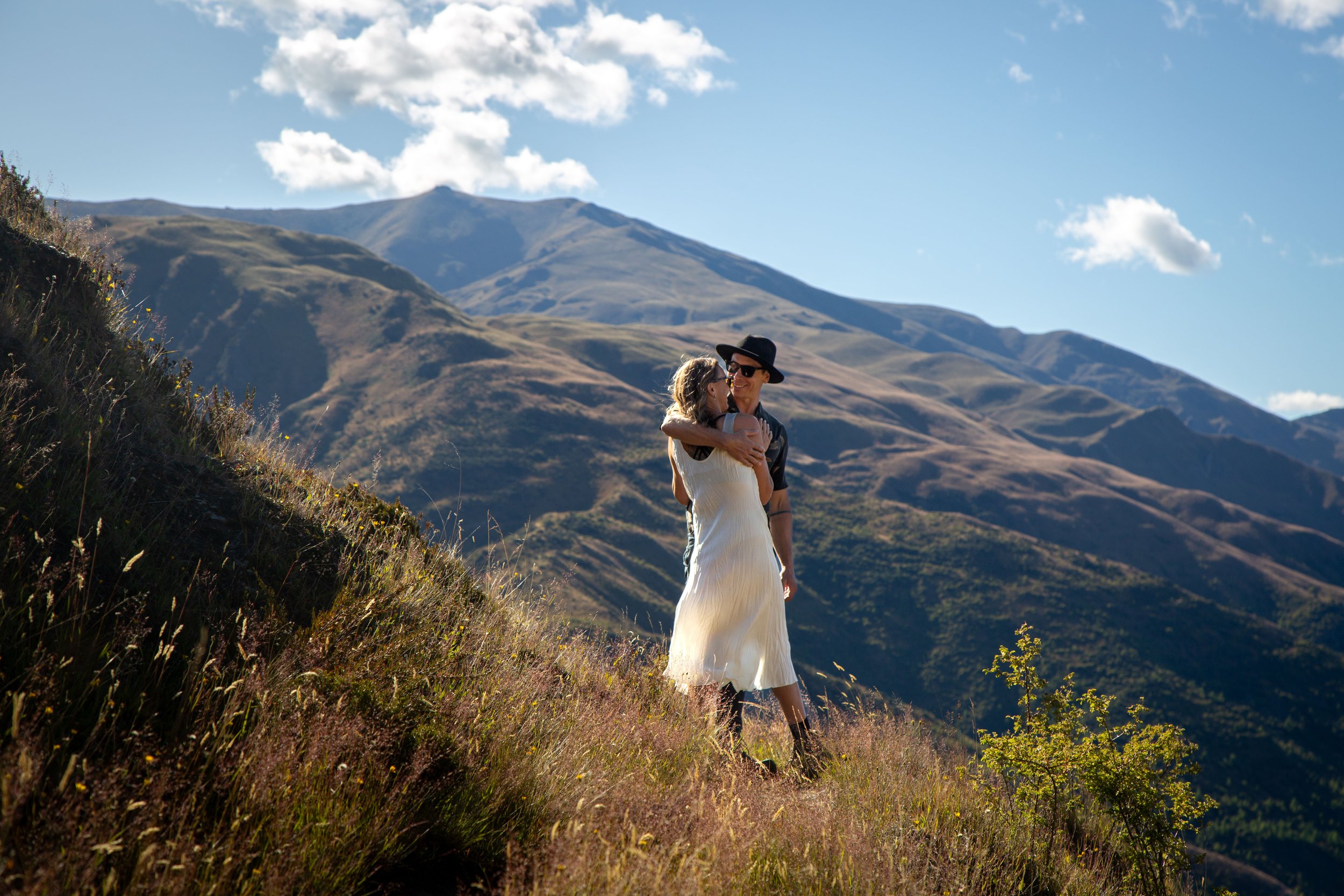 A couple hugging on a grassy hillside with mountains in the background under a clear sky.