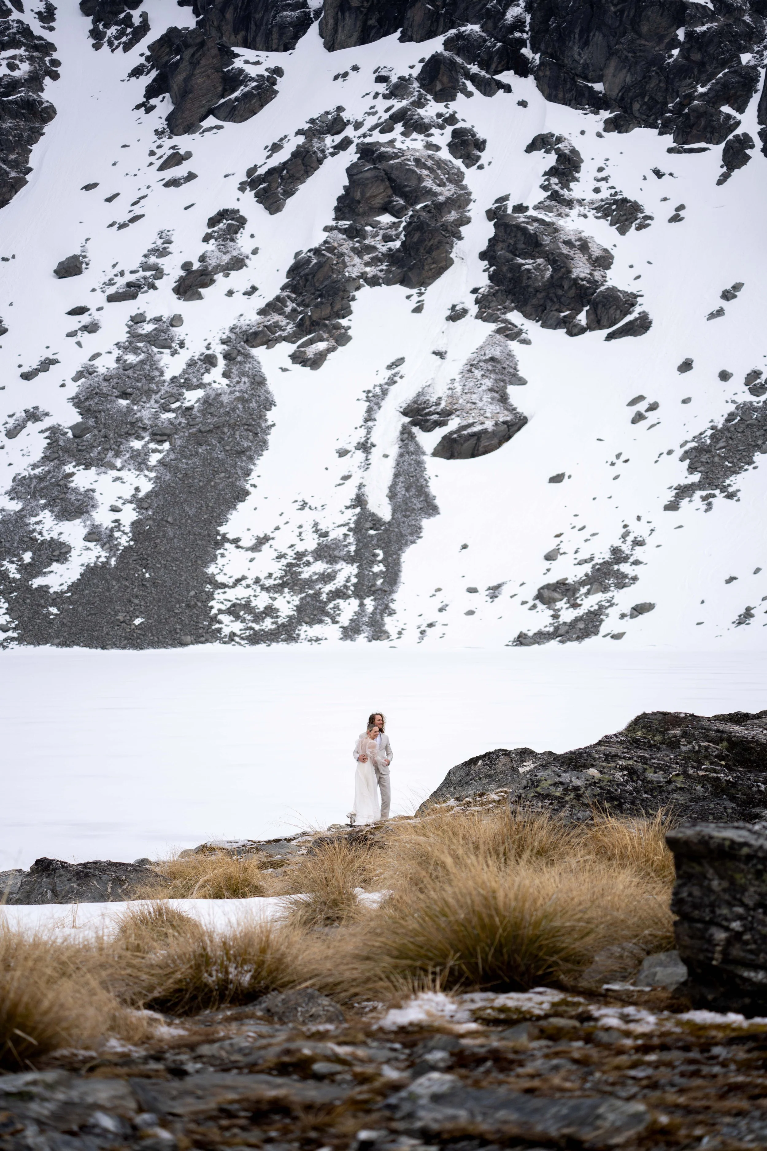 A couple standing on a rocky, grassy shoreline beside a frozen lake surrounded by snow-covered mountains.