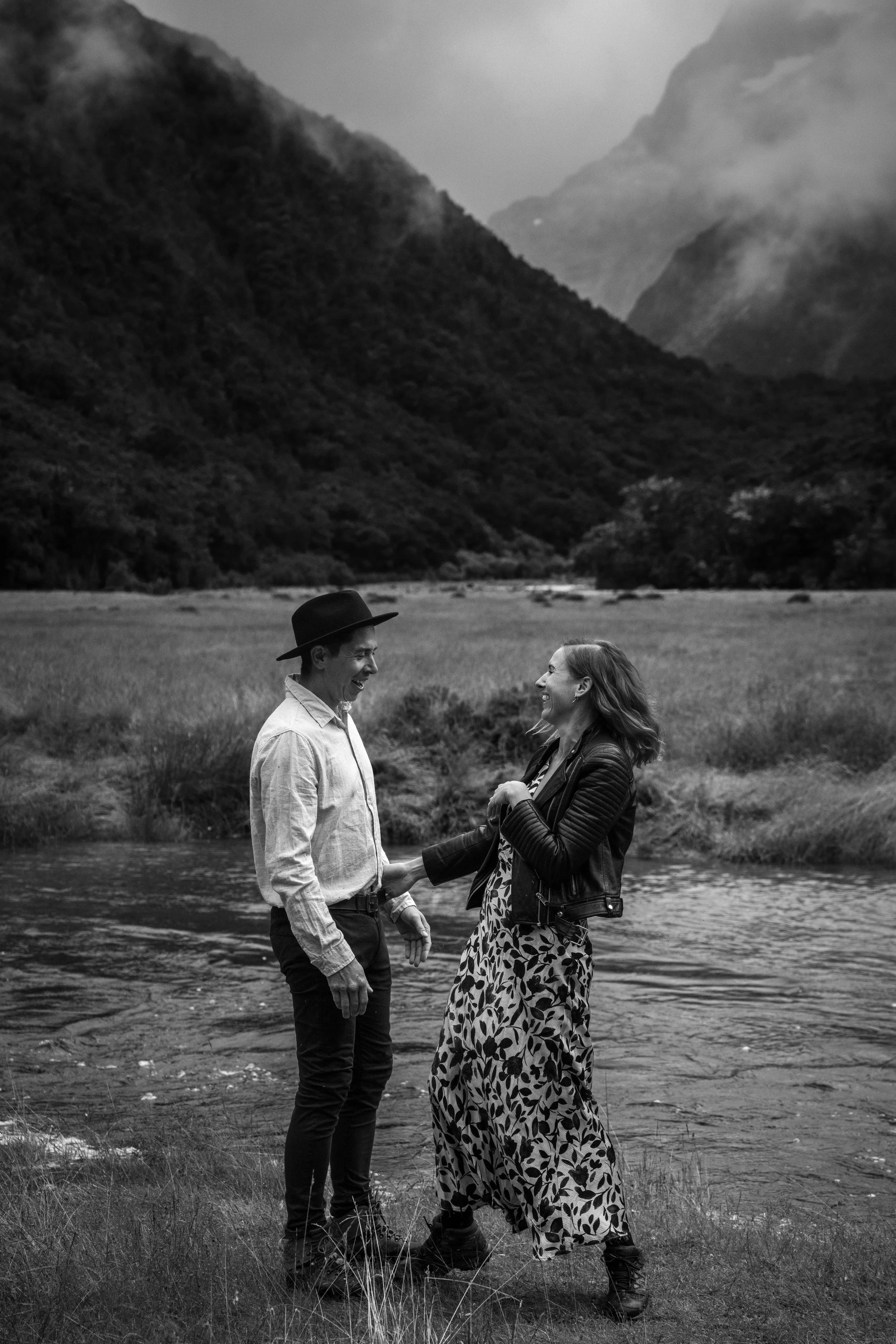 A black and white photo of a man and woman standing in a river, smiling and holding hands, with mountains and cloudy sky in the background.
