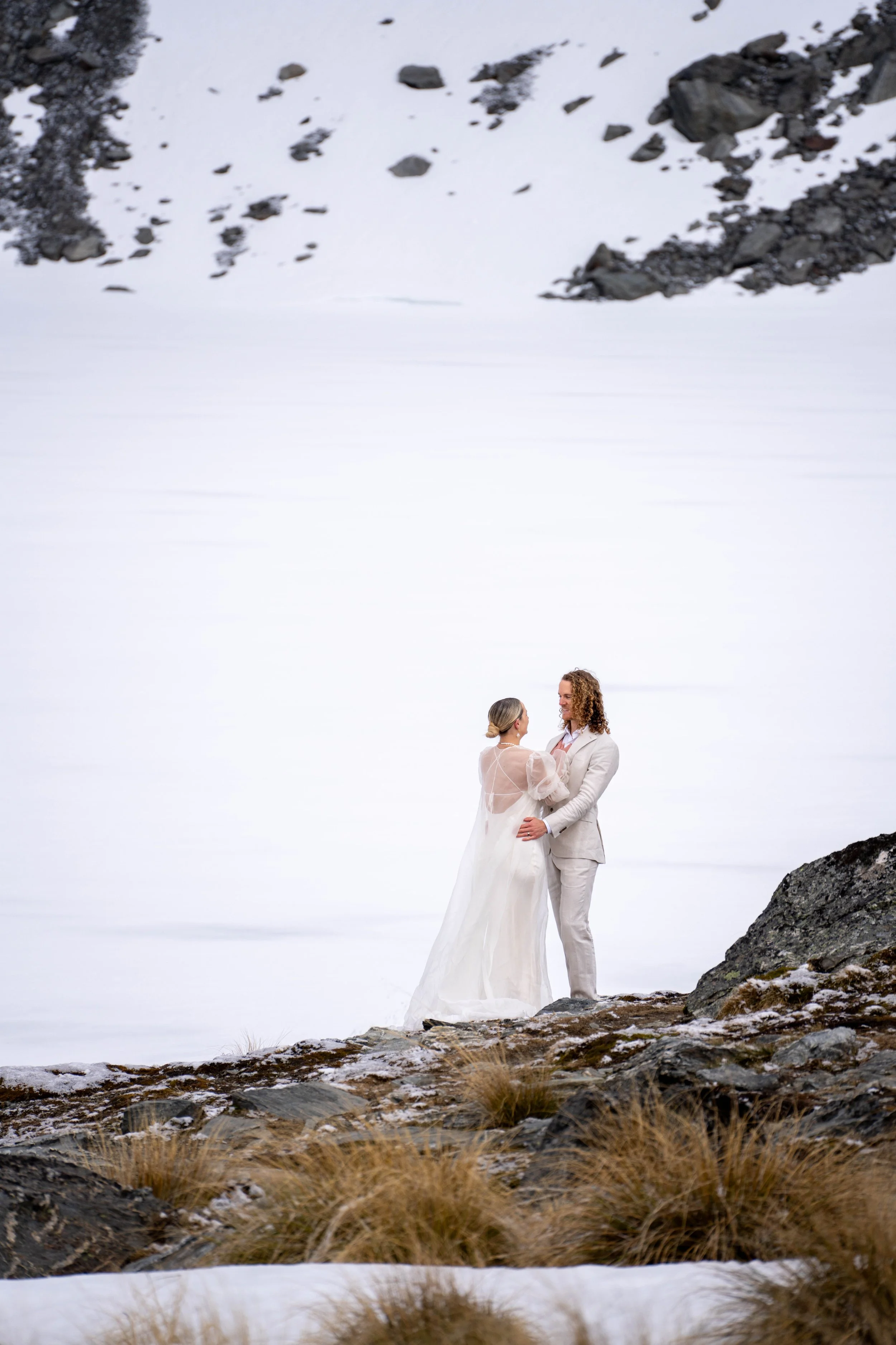 A couple in wedding attire standing on rocks near a snowy landscape, holding hands and gazing at each other.