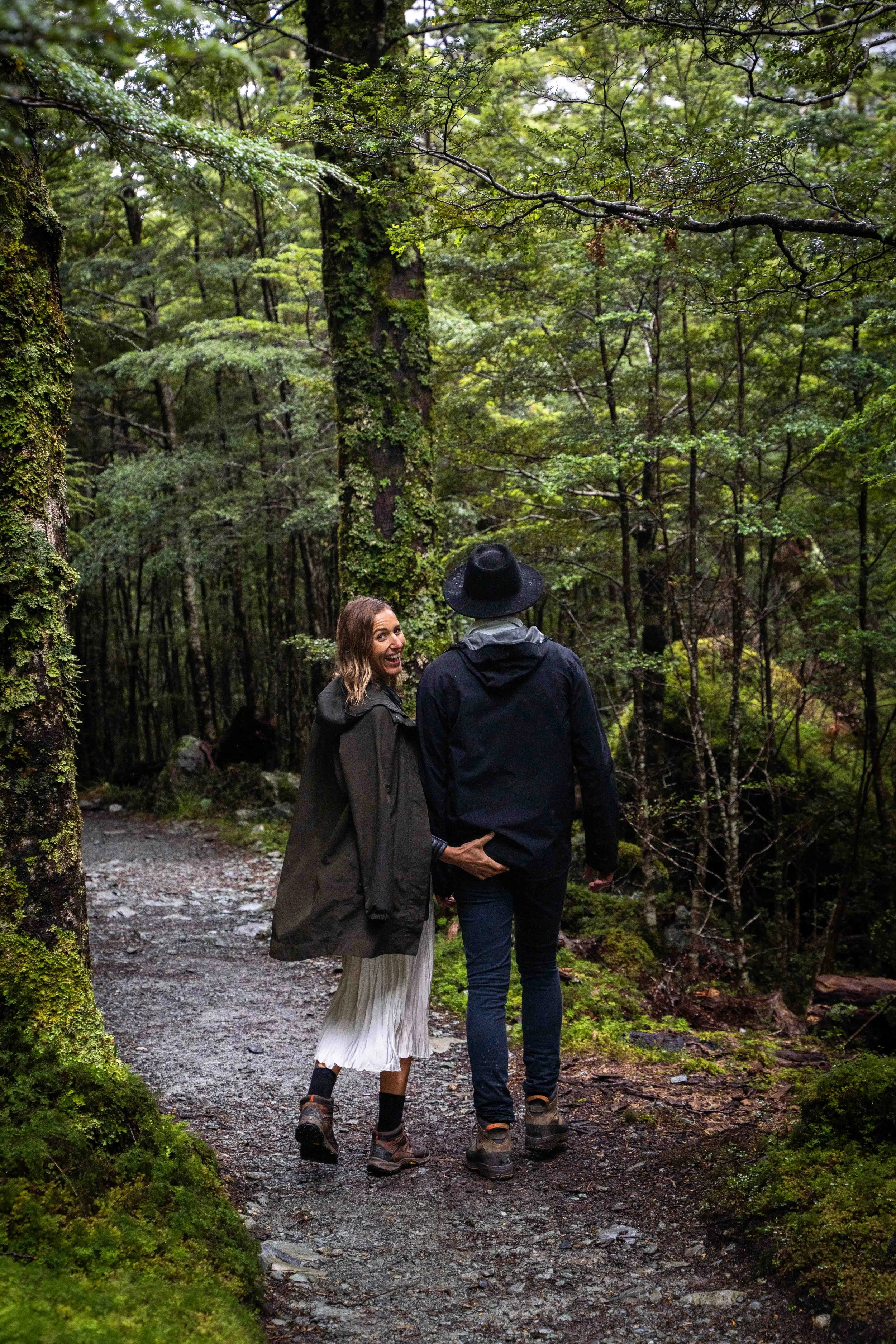 A woman and a man walk along a forest trail surrounded by lush green trees, with the woman smiling and looking back at the camera.