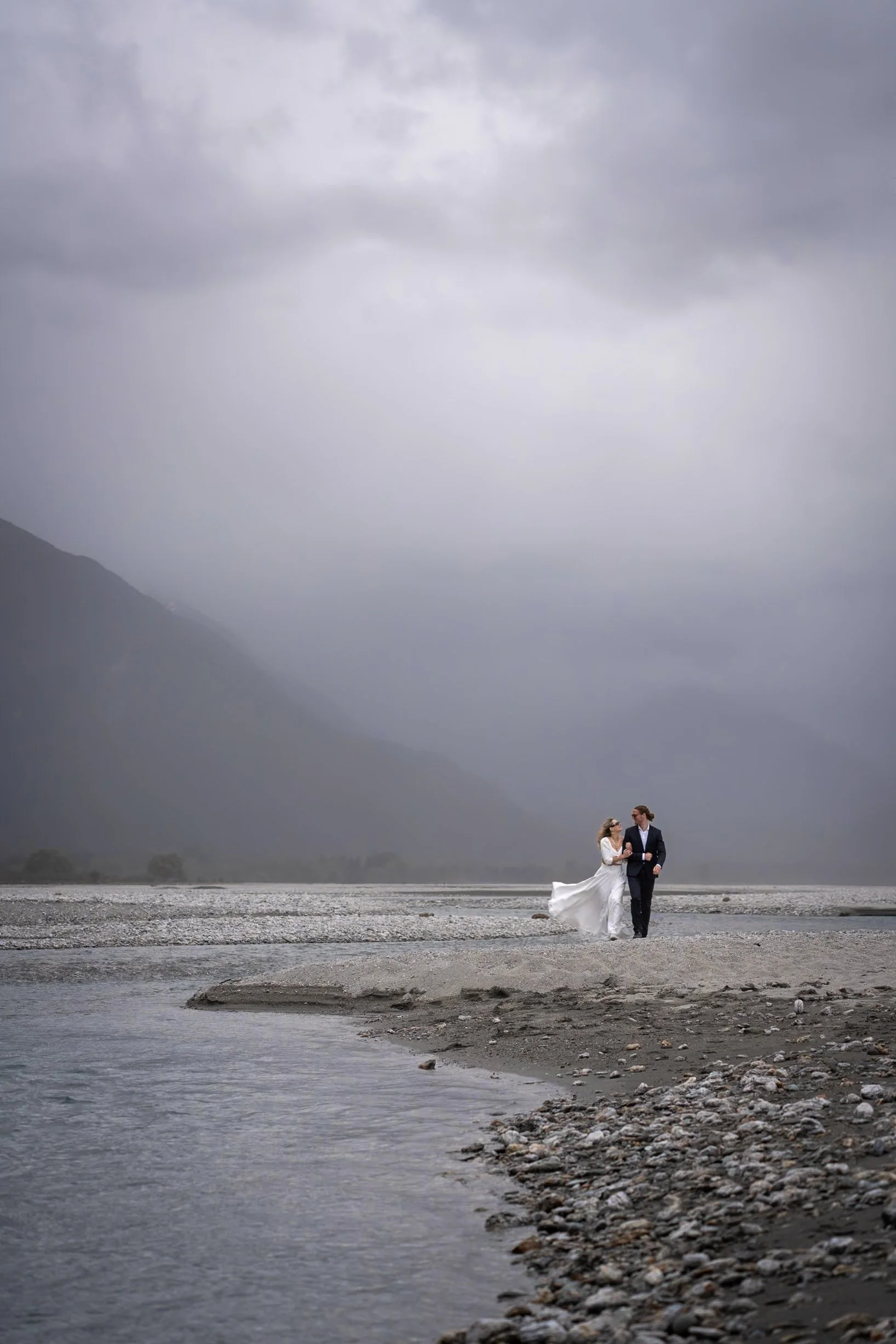 A bride and groom walking together on a rocky beach with a mountainous background and cloudy sky.