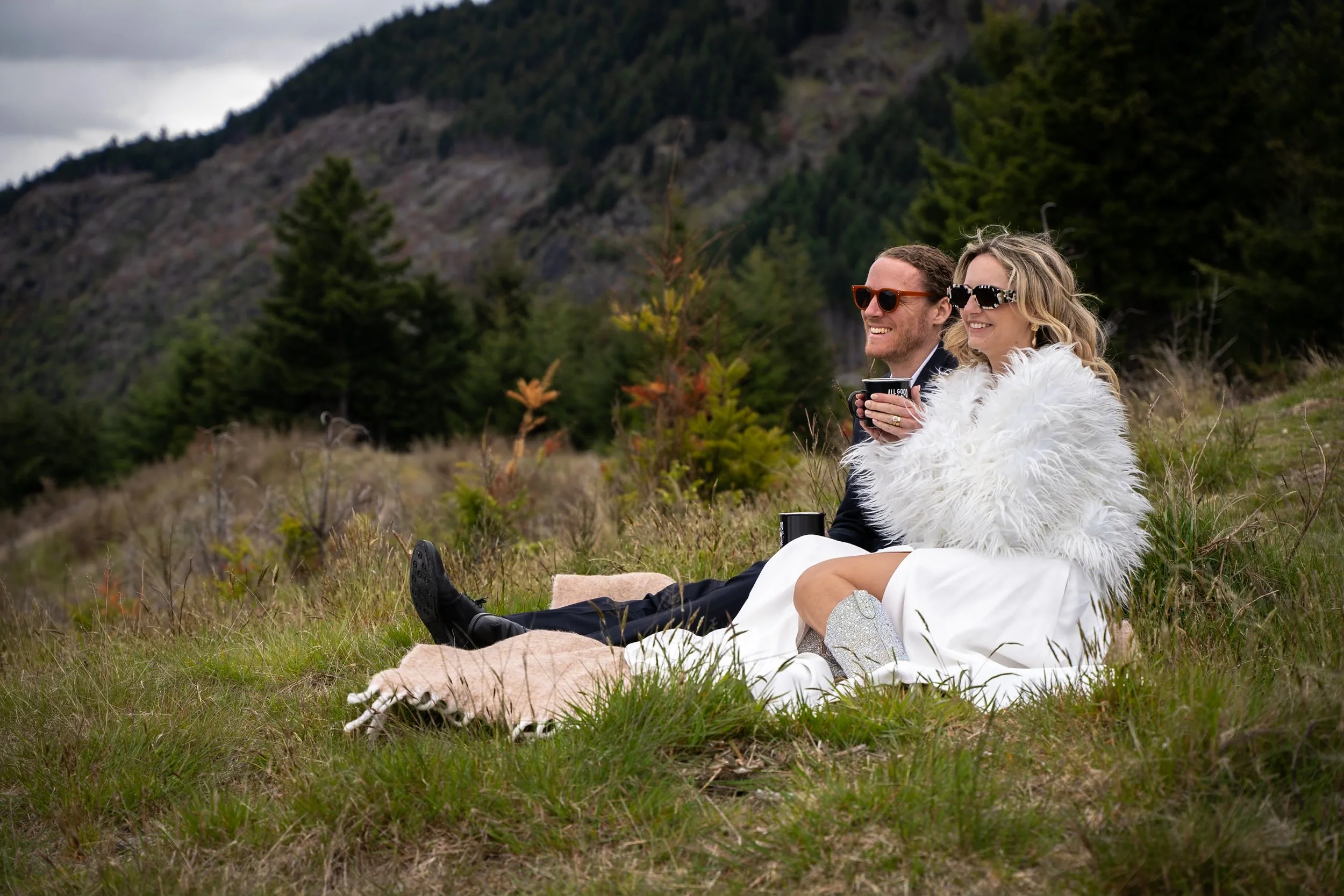 A happy couple in formal attire sitting on the grass in a scenic outdoor setting, enjoying drinks, with mountains and trees in the background.