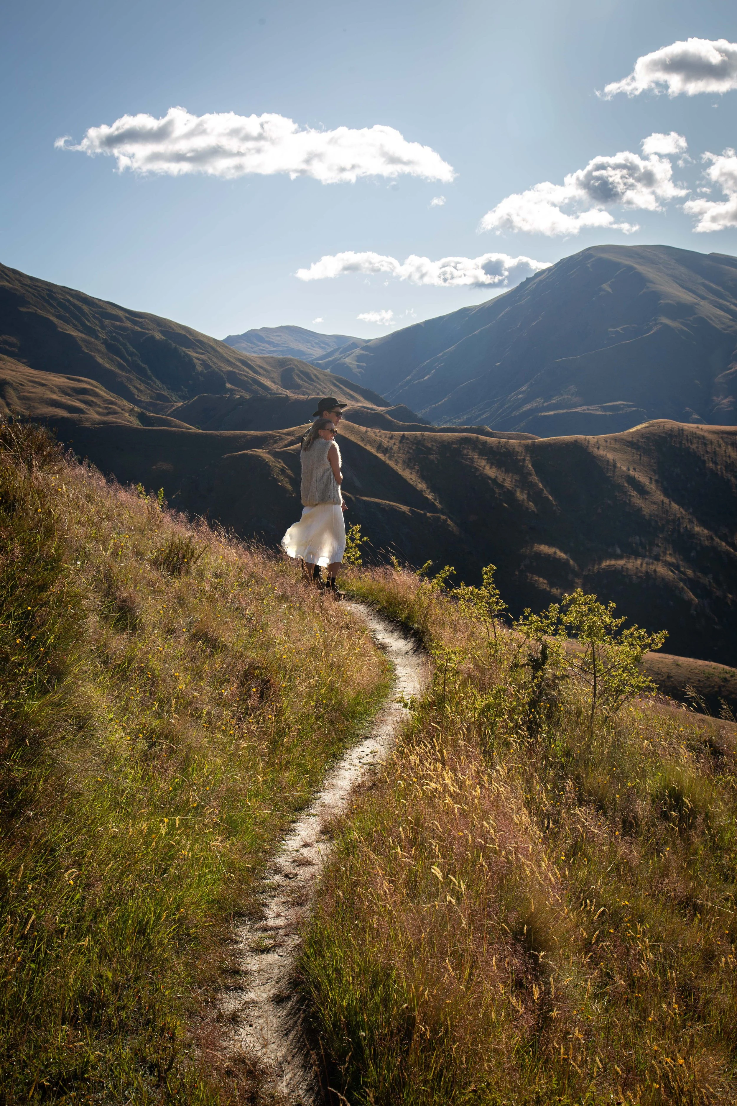 A woman and child walking along a narrow dirt trail on a hillside with mountains in the background under a partly cloudy sky.