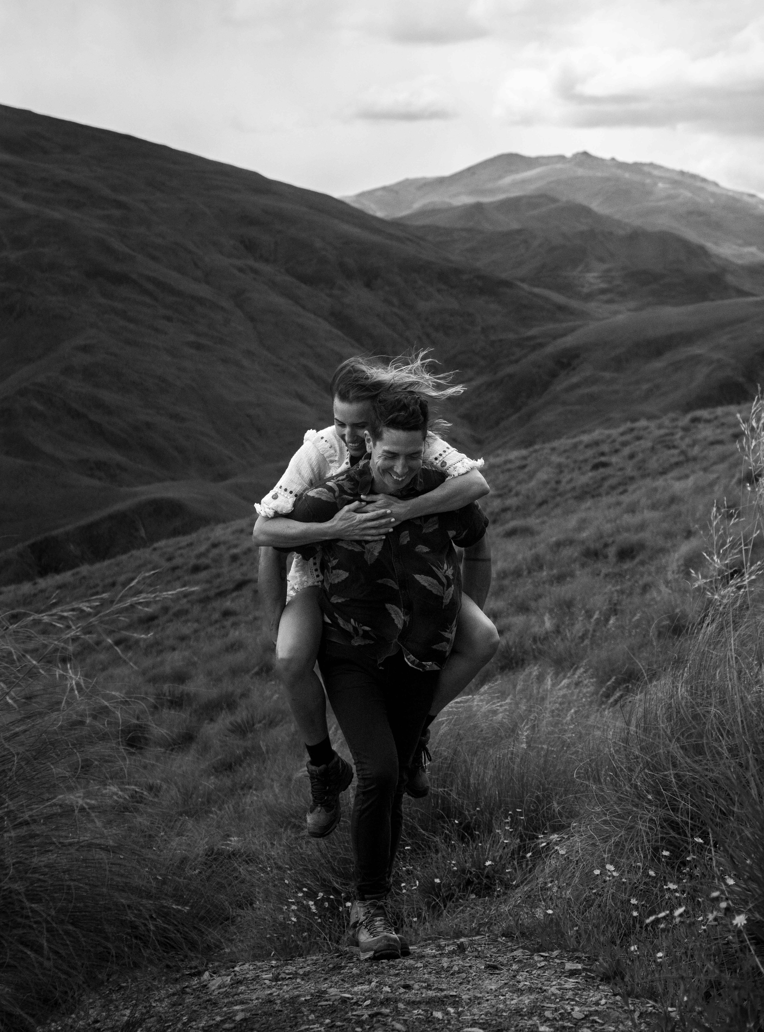 A black and white photo of a man carrying a woman on his back on a mountain trail with a scenic background of rolling hills and mountains.