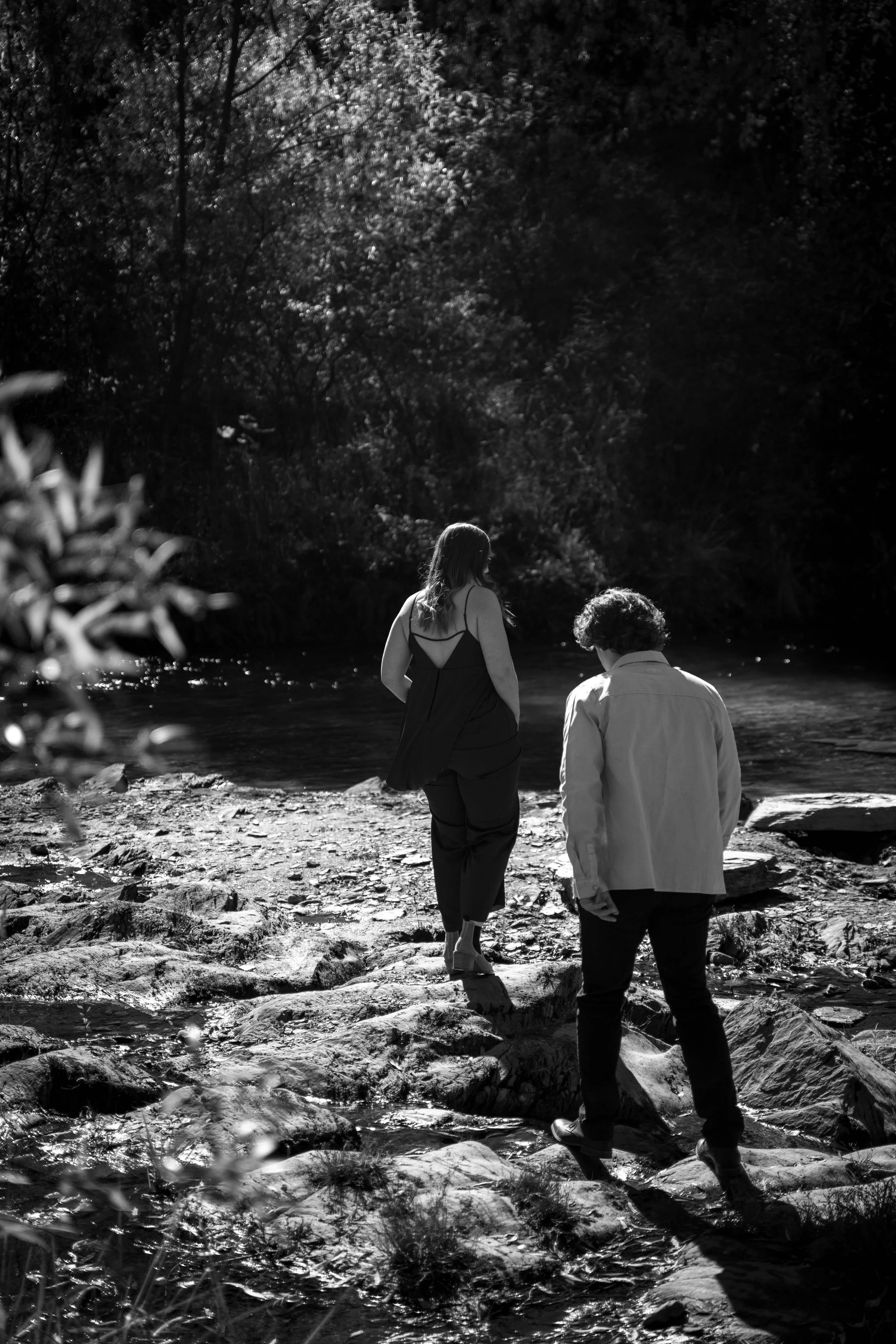 A black and white photo of two people walking across rocks near a stream or river in a wooded area, with trees and bushes in the background.