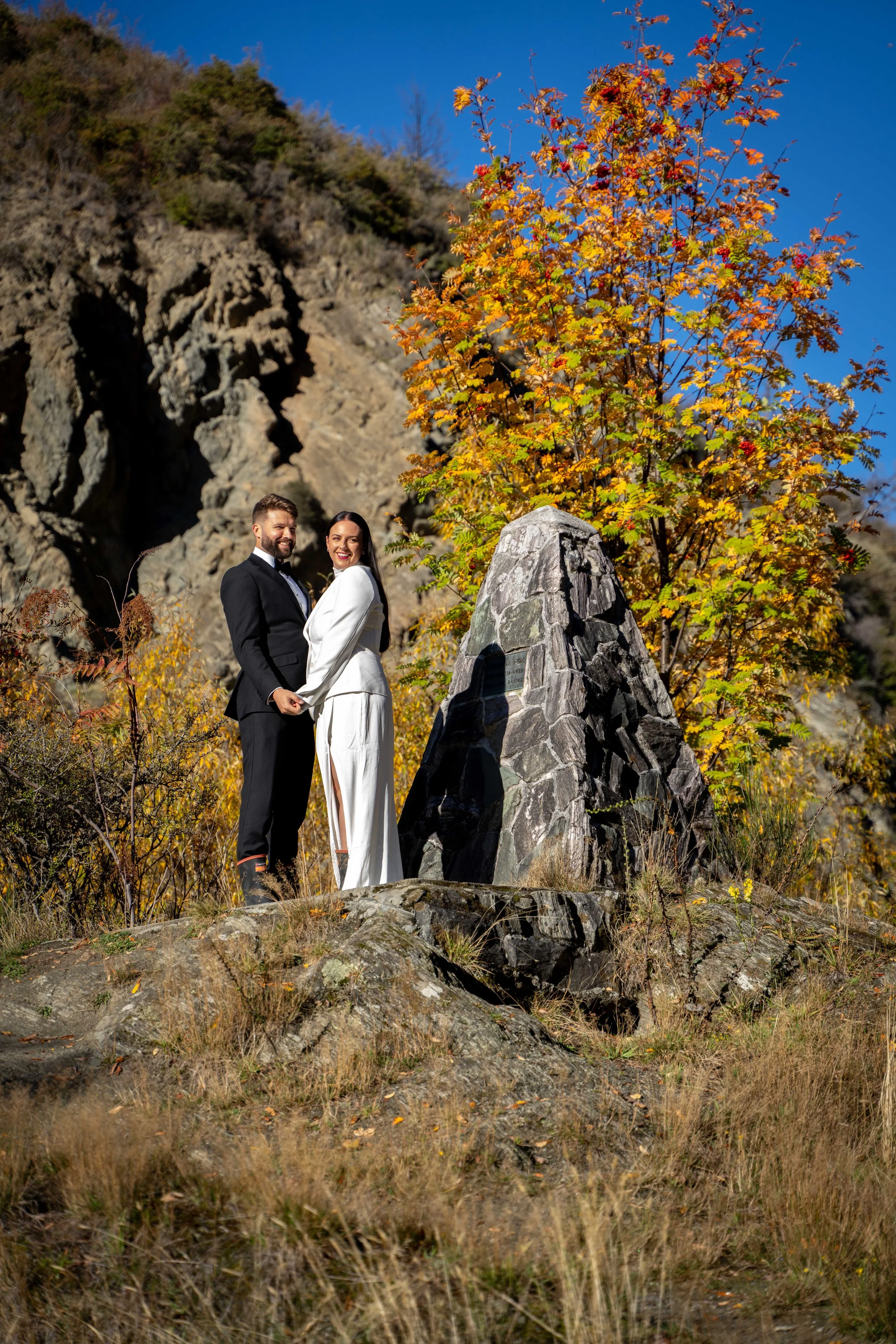 A couple in wedding attire holding hands and smiling outdoors in front of a rock formation and autumn-colored trees.