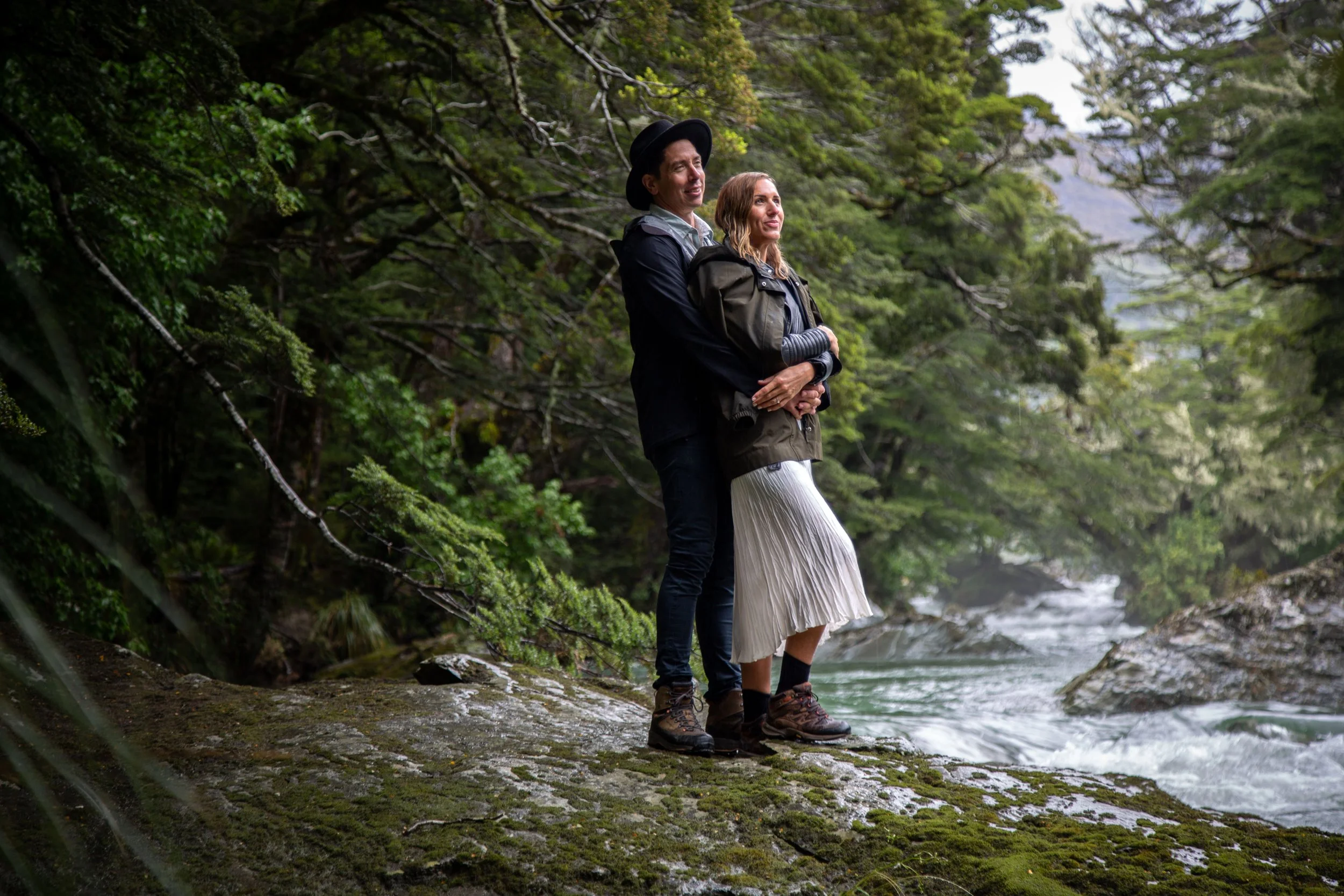 A couple stands on a mossy rock by a river in a lush forest, embracing and looking into the distance.