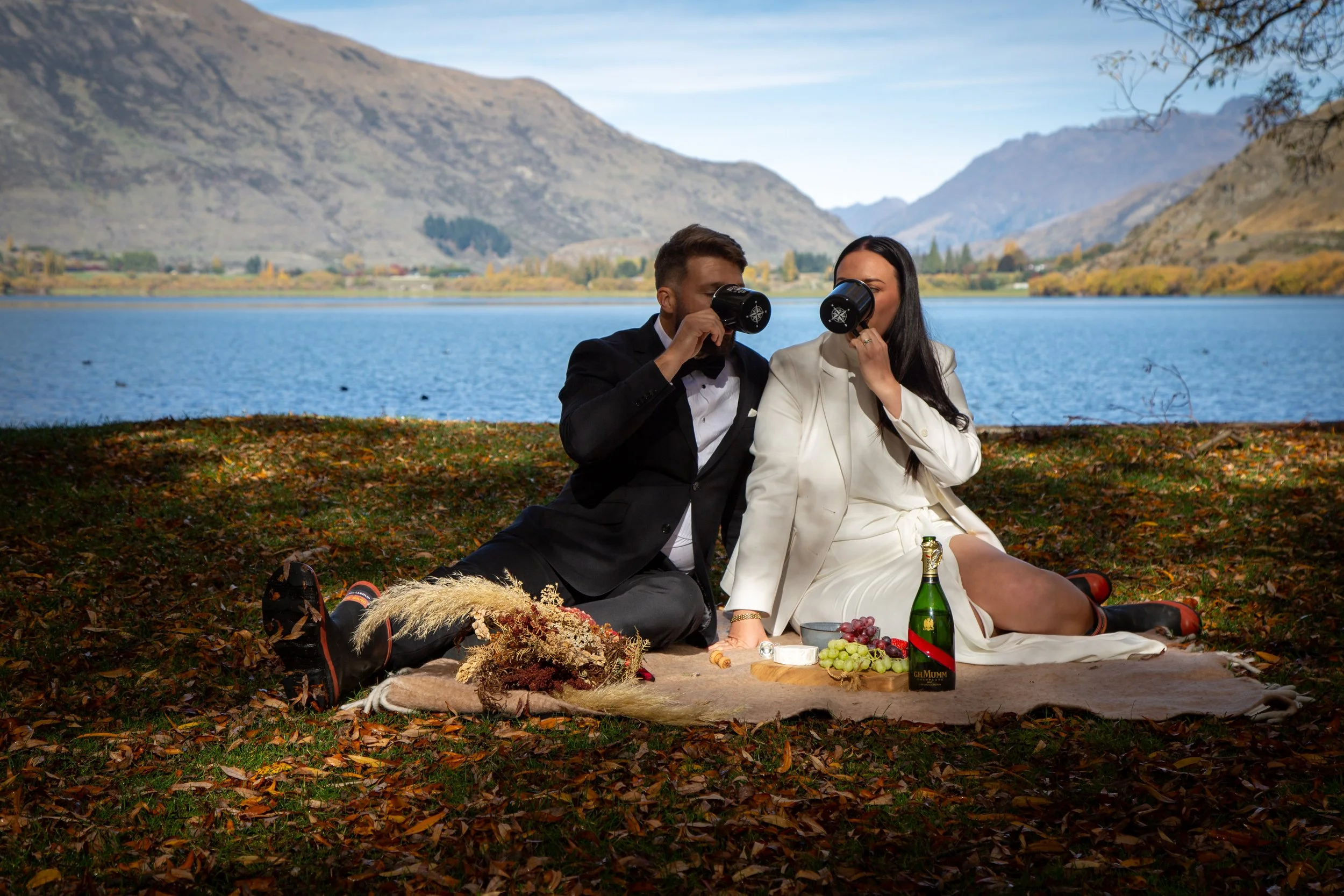 A couple in formal attire having a picnic outdoors near a lake with mountains in the background, drinking from black mugs.
