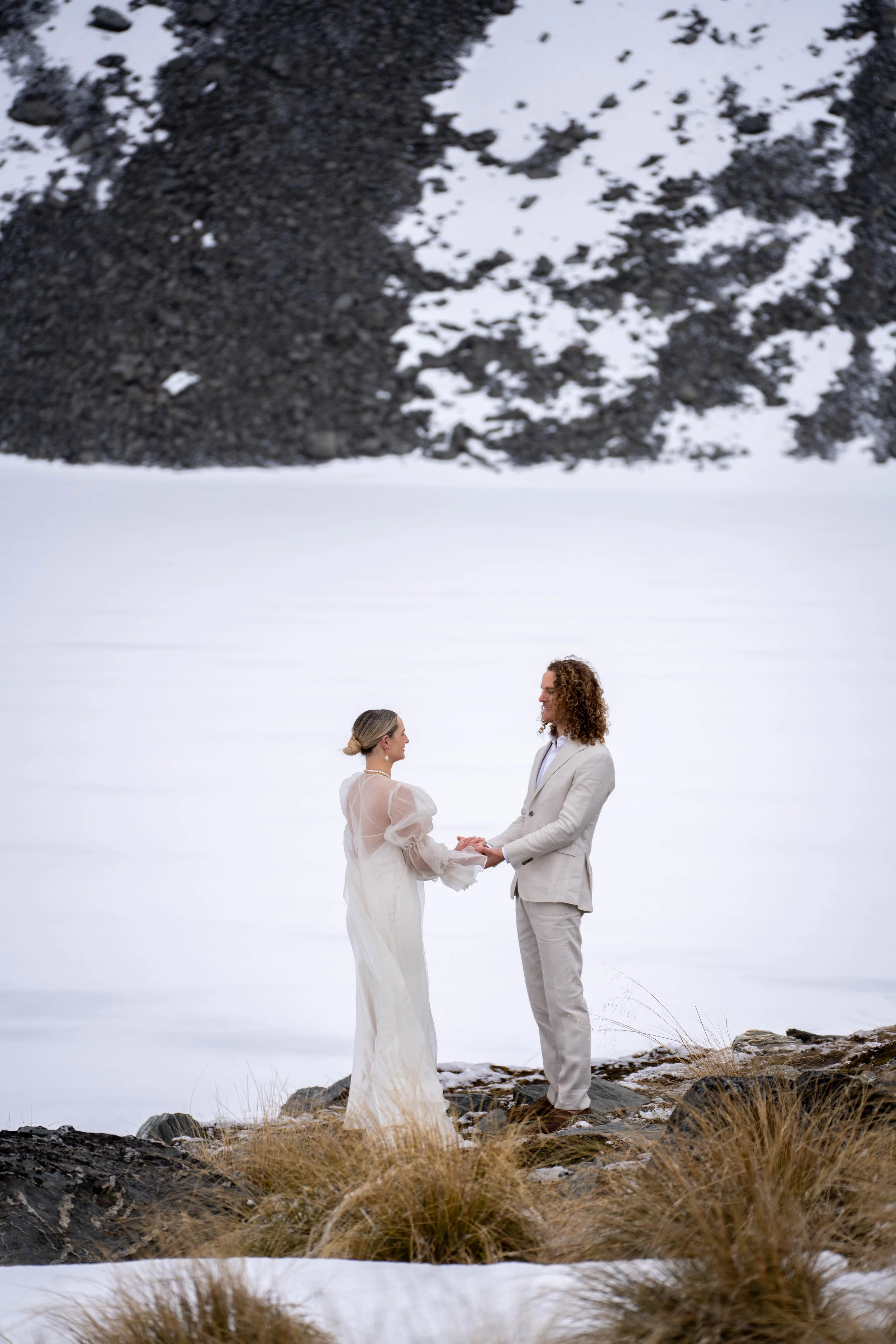 A bride and groom holding hands and facing each other on a snowy landscape, with rocky terrain and dried grass in the foreground.