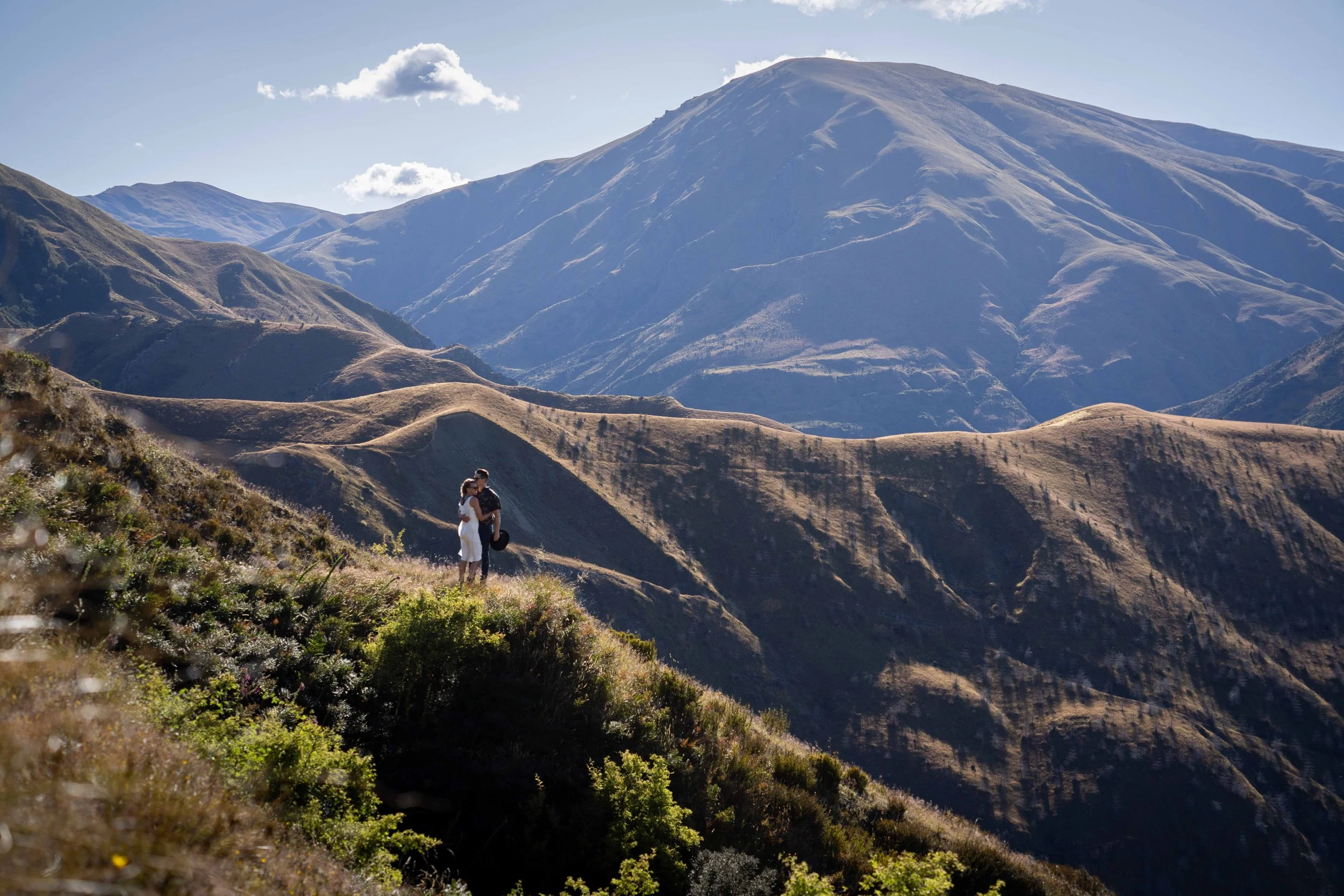 A couple standing on a hillside in a mountainous landscape with rolling hills and large mountains in the background under a partly cloudy sky.