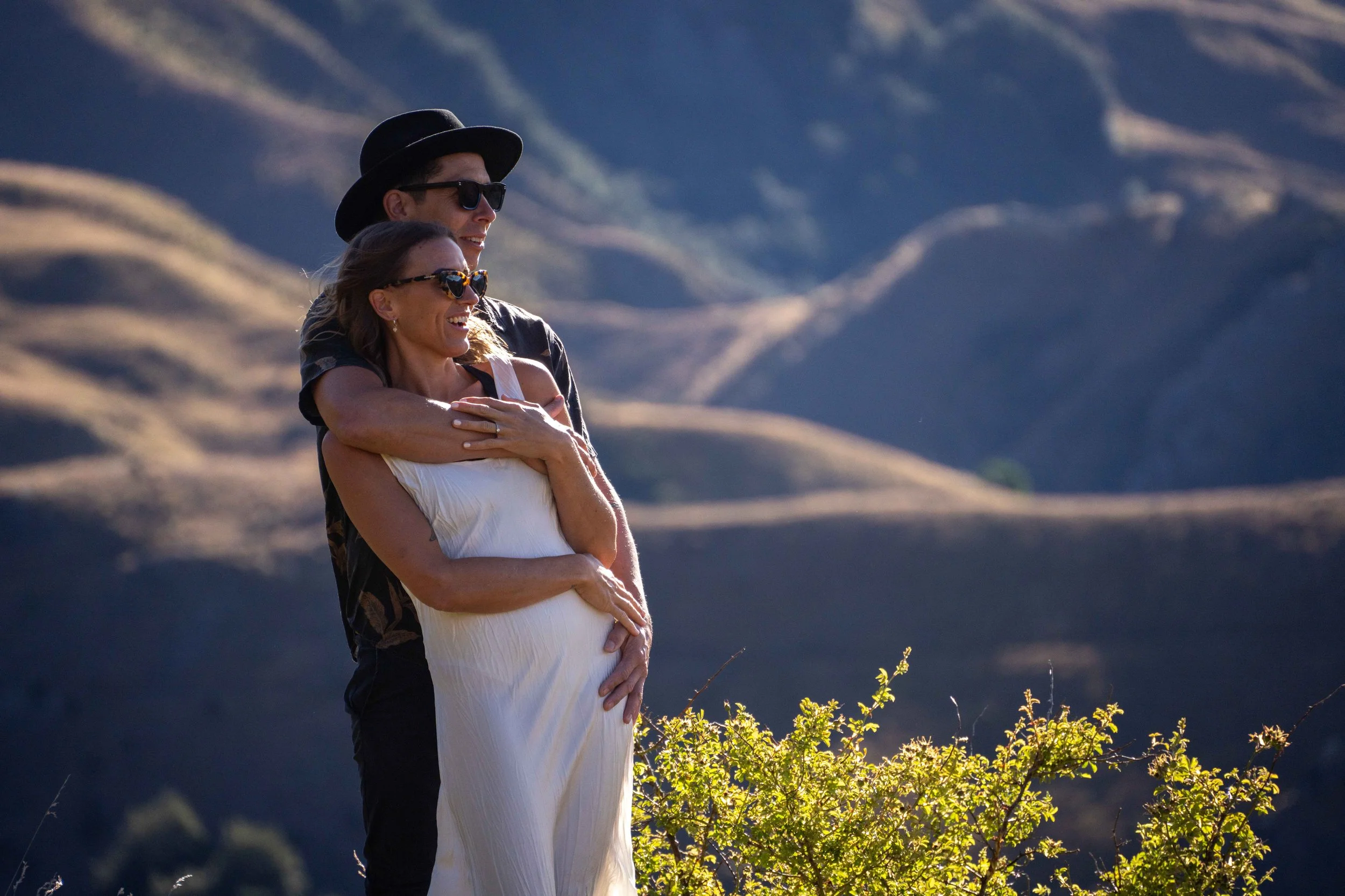 A couple stands outdoors admiring the view in a mountainous area, with the man hugging the woman from behind, both smiling and wearing sunglasses.