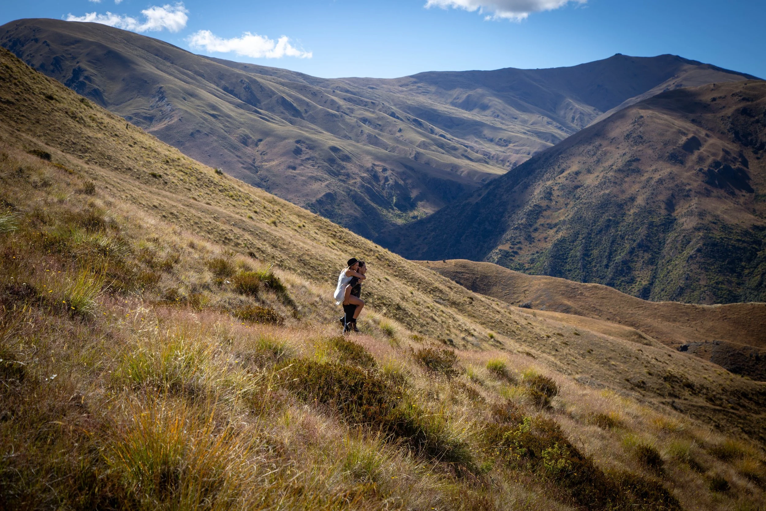 A couple hiking in a mountainous landscape with grassy slopes and rolling hills under a blue sky.