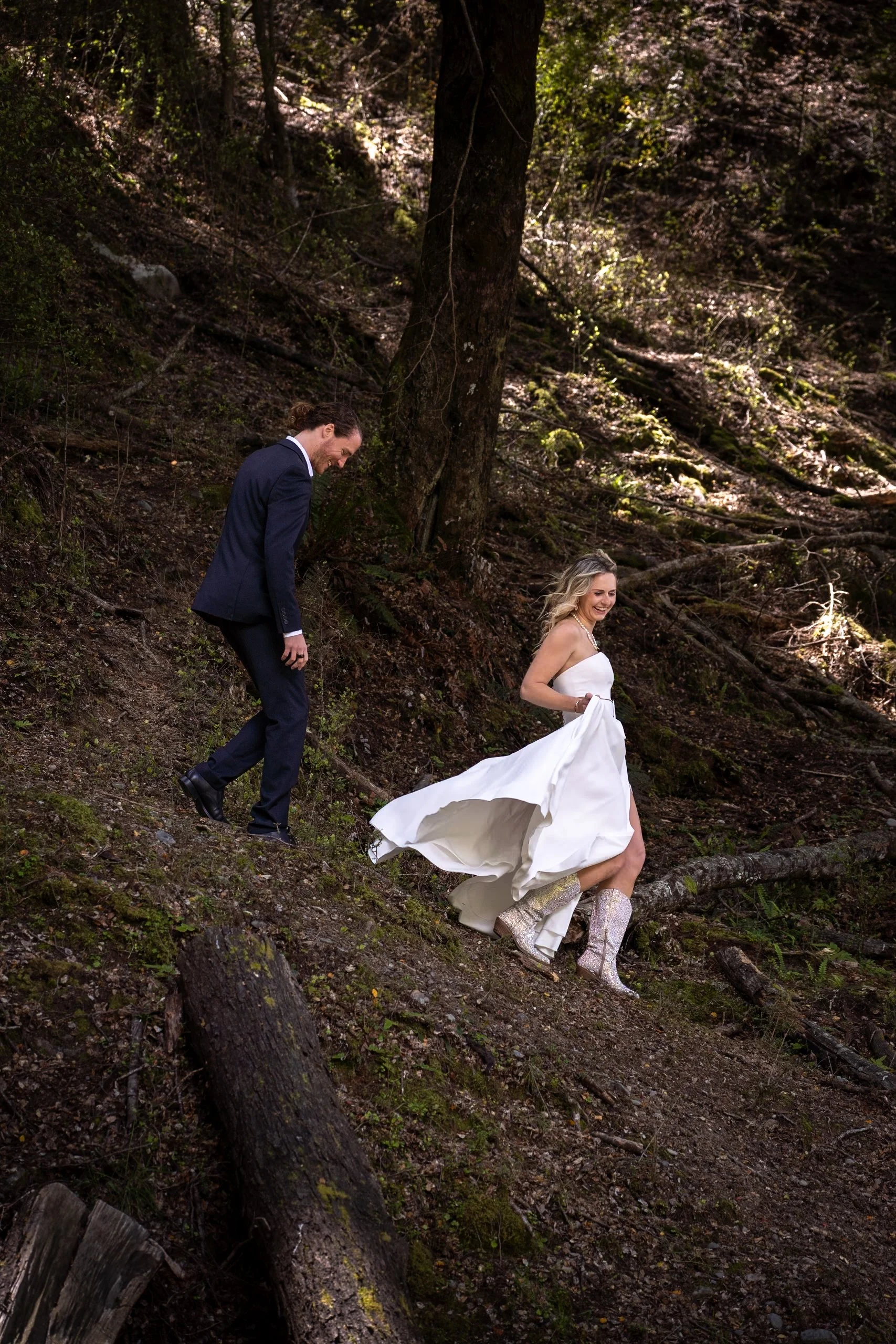 A bride in a white wedding dress and glittery cowboy boots and a groom in a dark suit hiking down a rocky wooded trail, smiling and enjoying themselves.