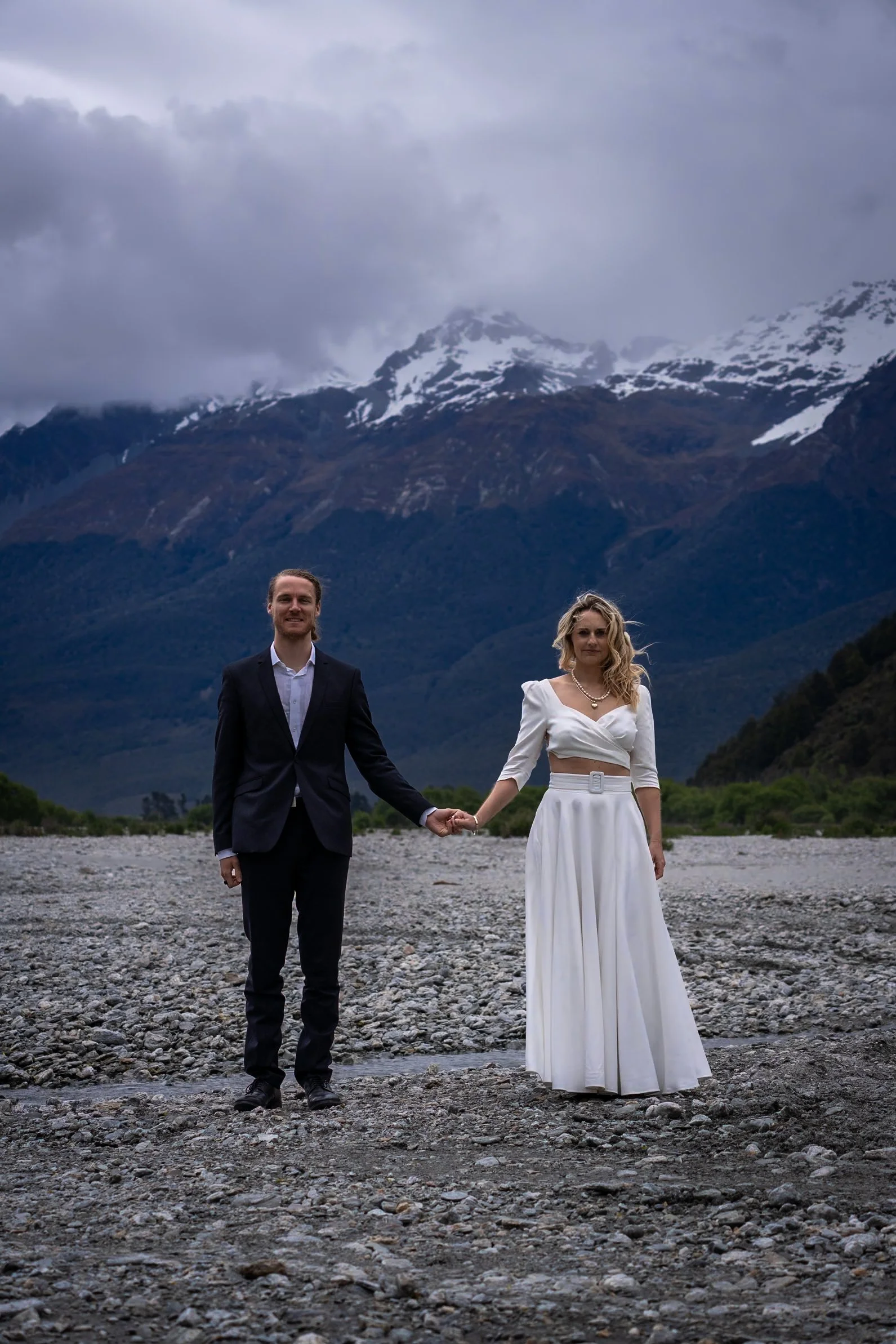 A man and woman in formal attire holding hands in a rocky outdoor setting with mountains and snow in the background.