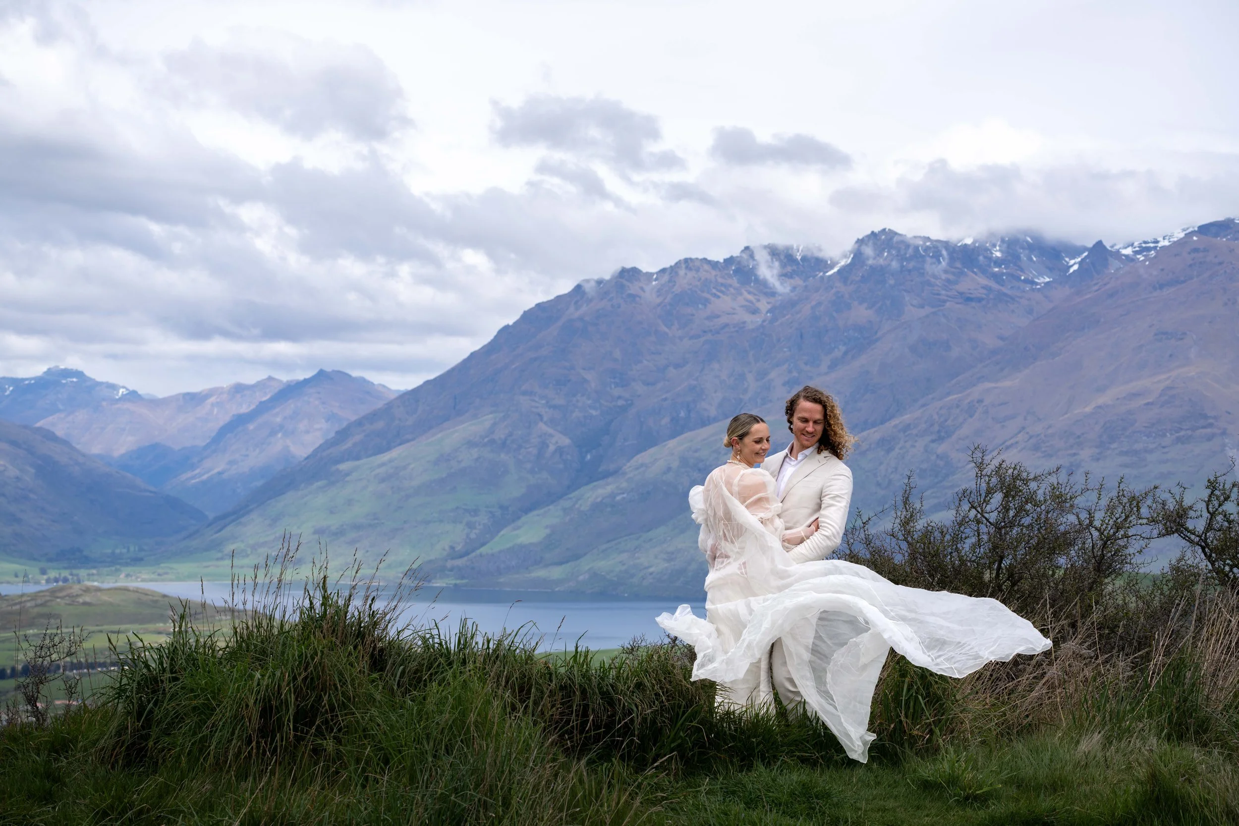 A couple in wedding attire sitting together outdoors on grass with a mountain landscape in the background, including a lake and cloudy sky.