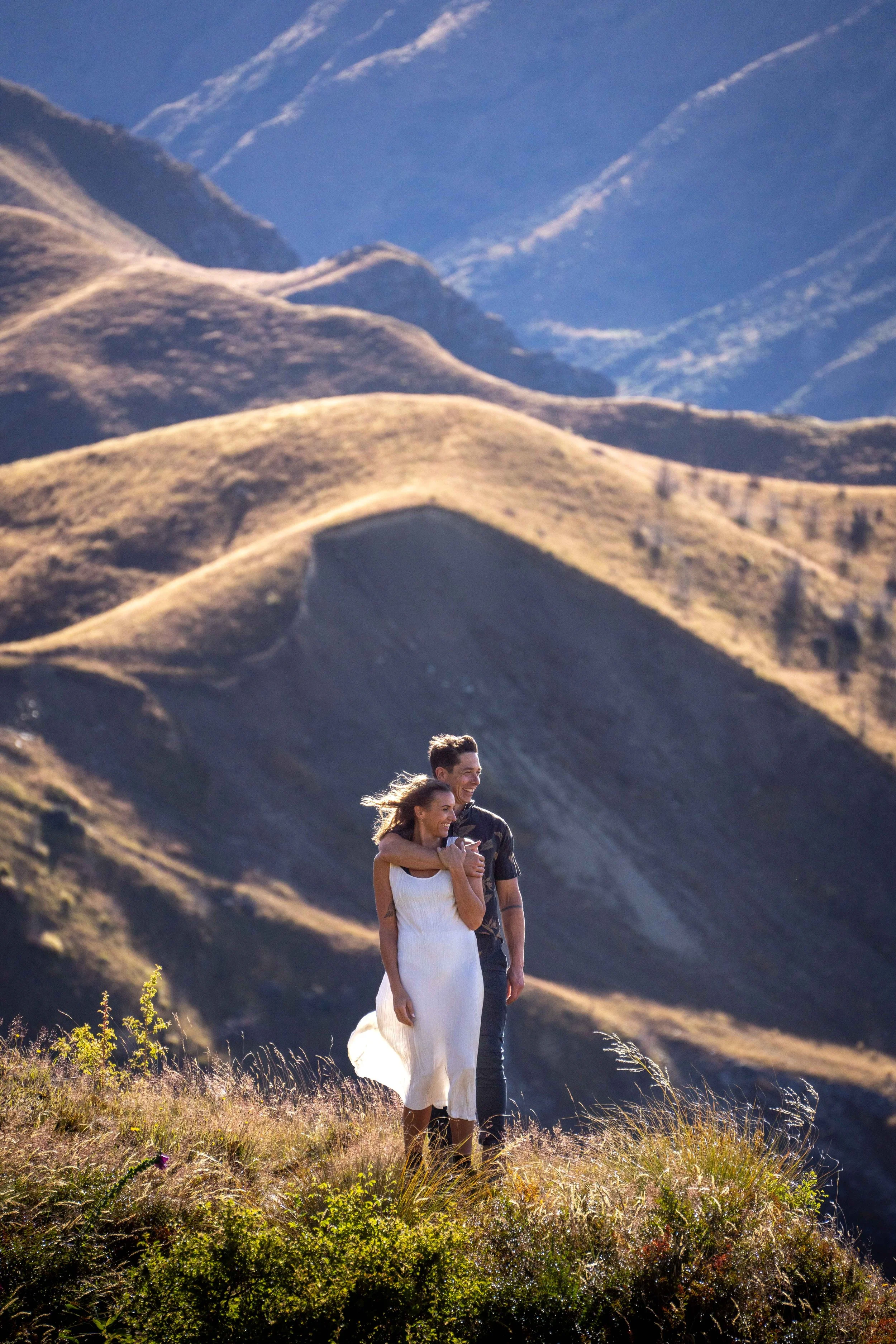 A couple stands together on a grassy hill with mountainous terrain in the background, enjoying a moment outdoors.