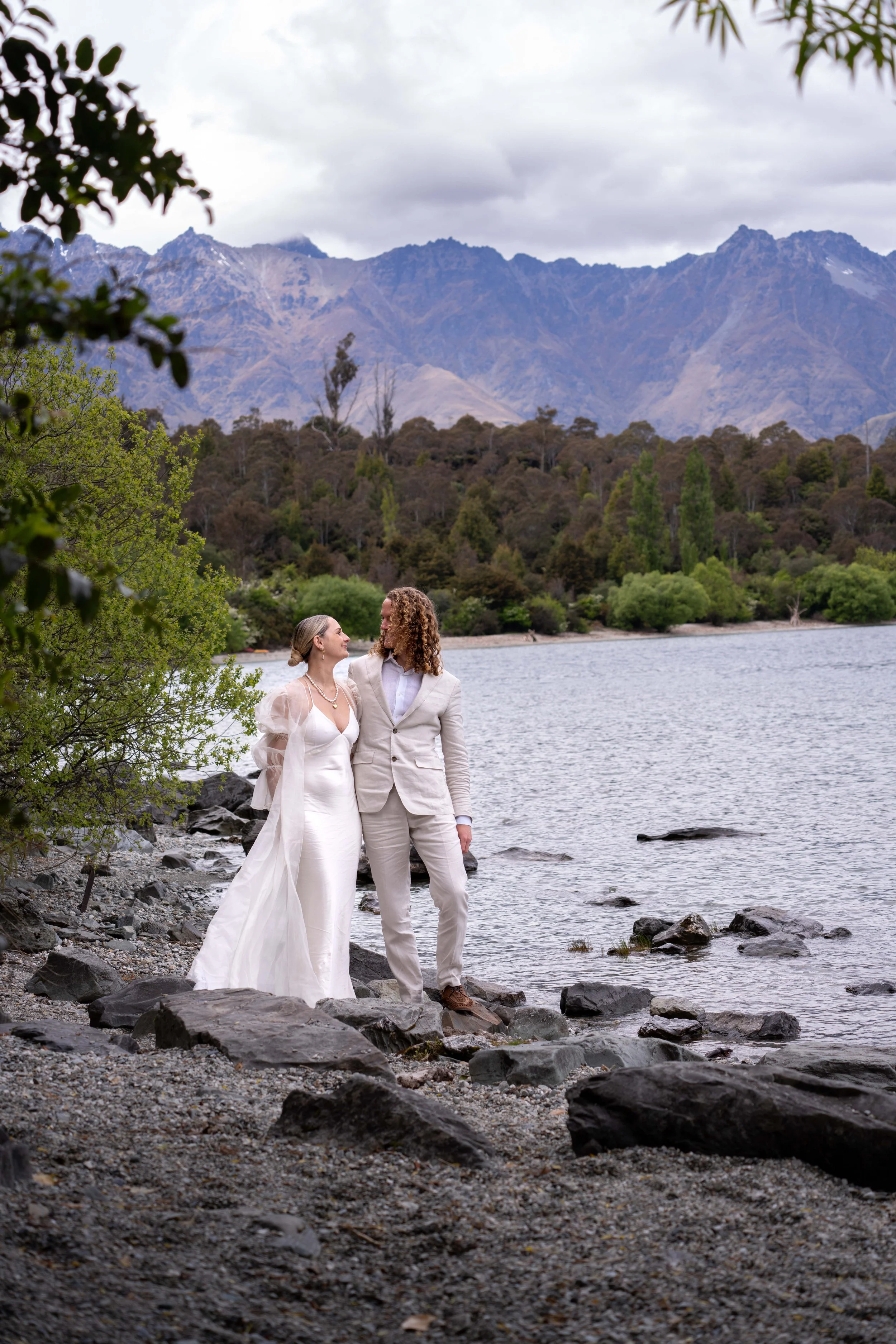 A couple dressed in white wedding attire standing on rocky beach by lake, surrounded by trees and mountain landscape under cloudy sky.