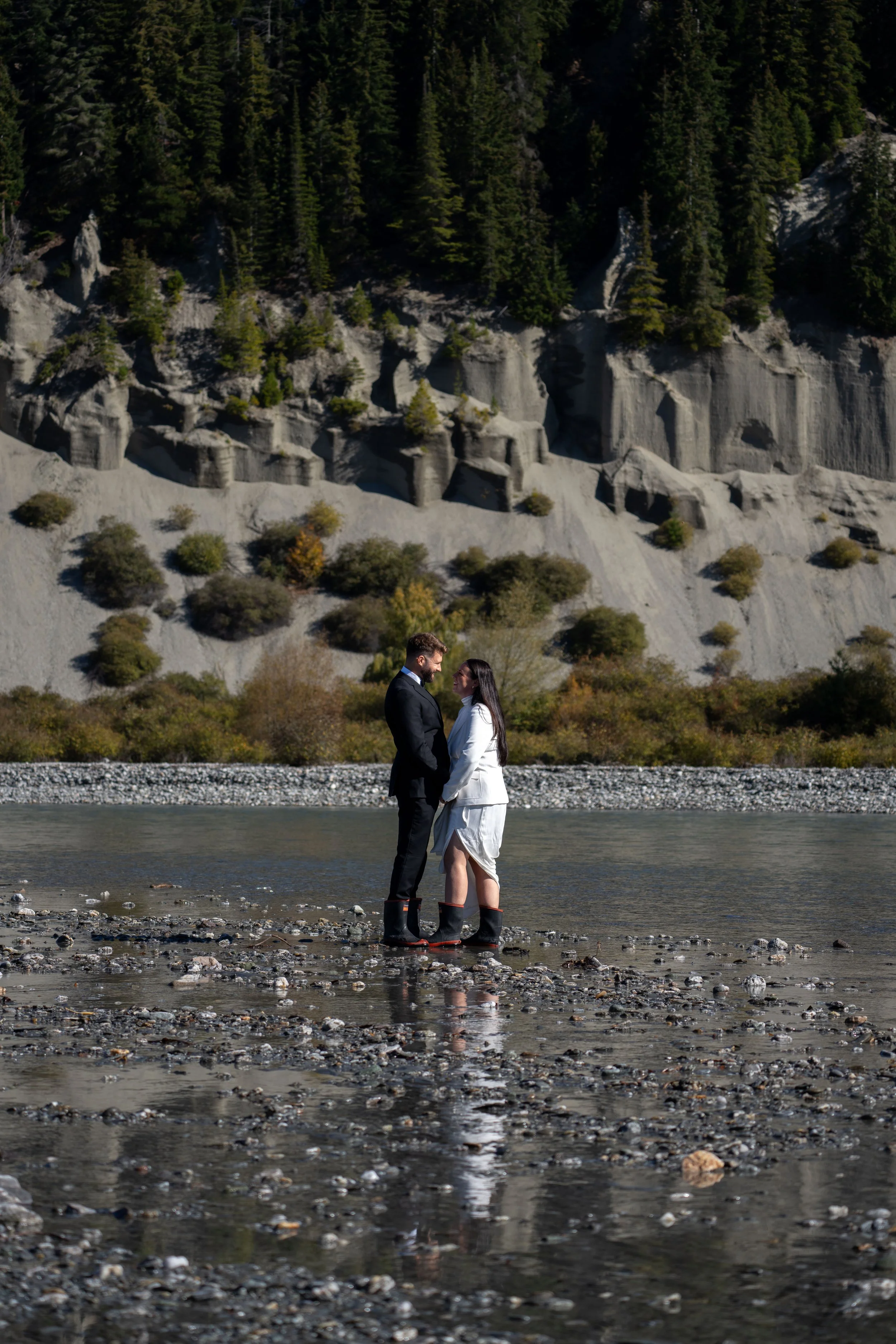 A couple stands in a shallow river, holding hands and looking at each other, with tall trees and rocky cliffs in the background.