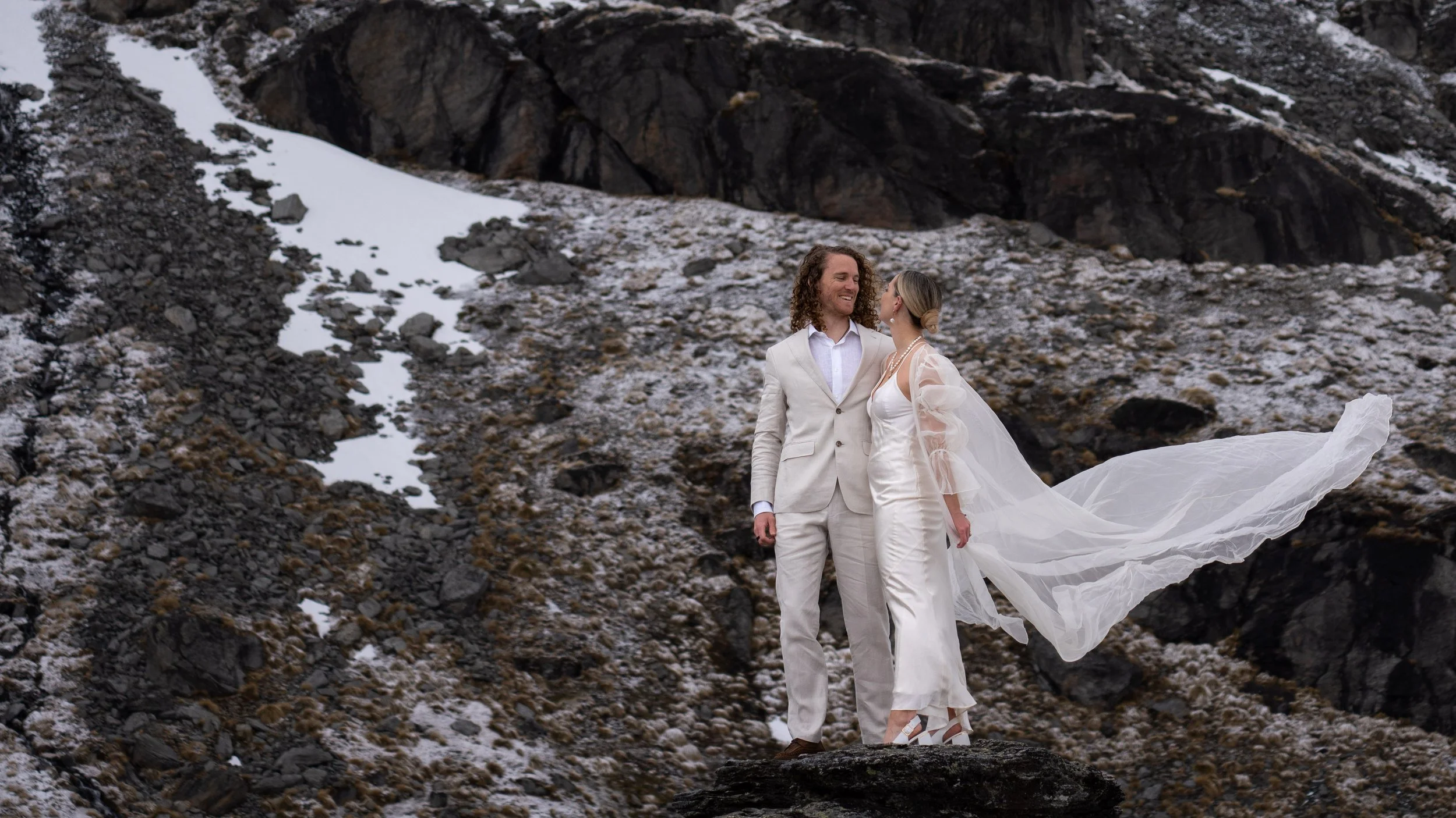 A couple dressed in wedding attire standing on a rock in a mountainous, rocky landscape with patches of snow. The groom has curly hair and wears a light-colored suit, while the bride has her hair in a bun and wears a white dress with a flowing veil.