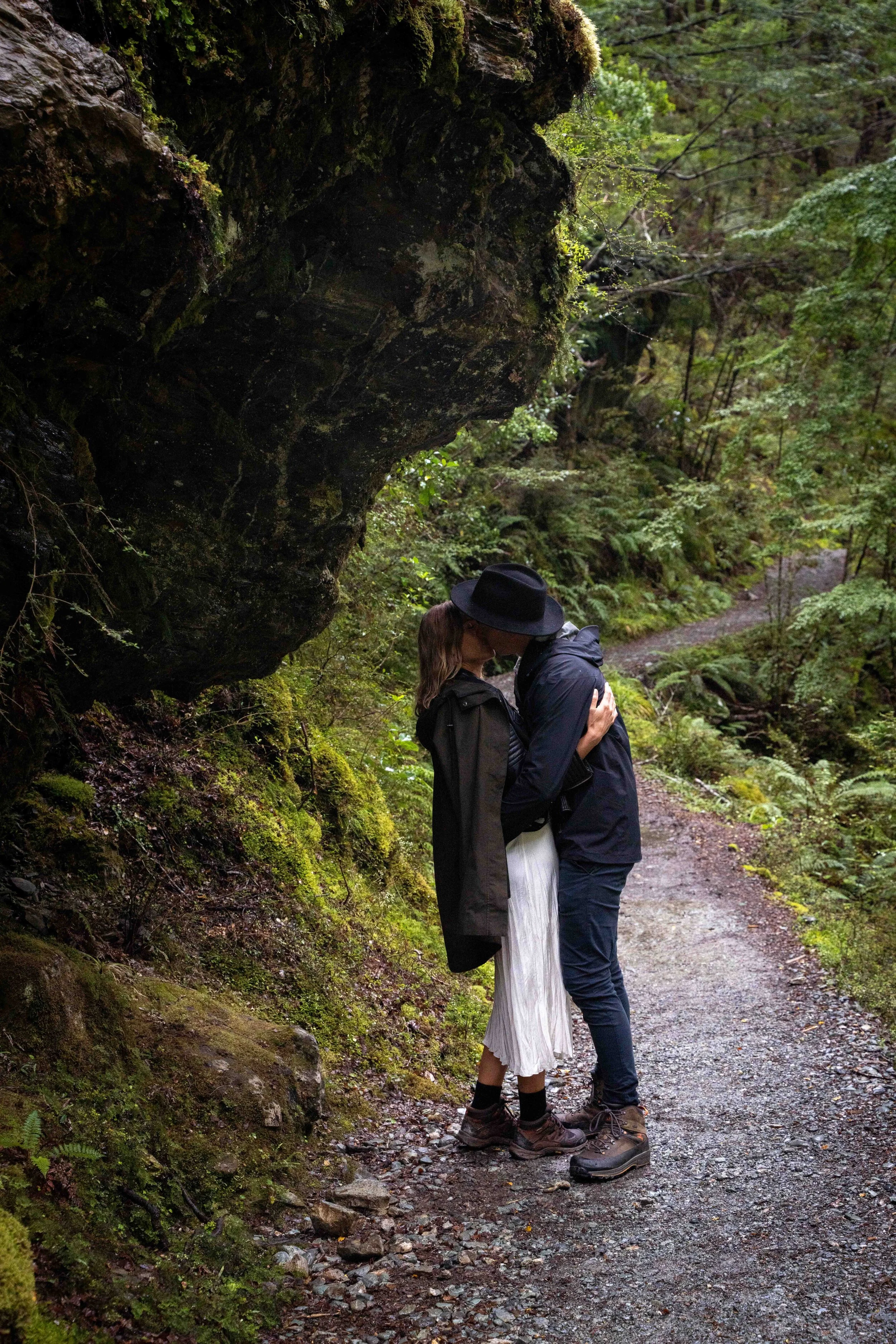 A couple kissing on a forest trail, surrounded by green moss and trees.