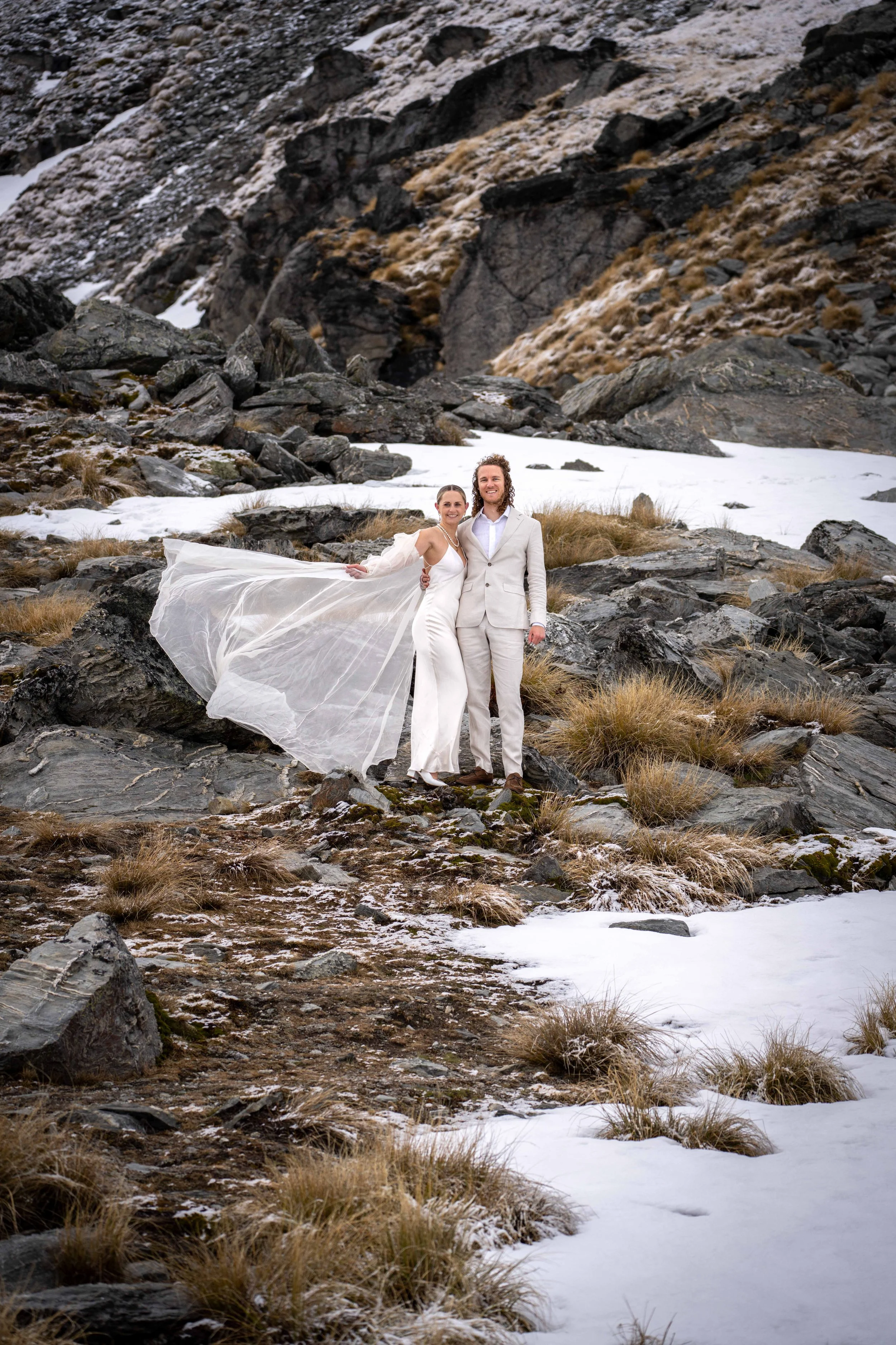 A bride and groom in wedding attire standing on rocky terrain with patches of snow and dry grass, mountains in the background.