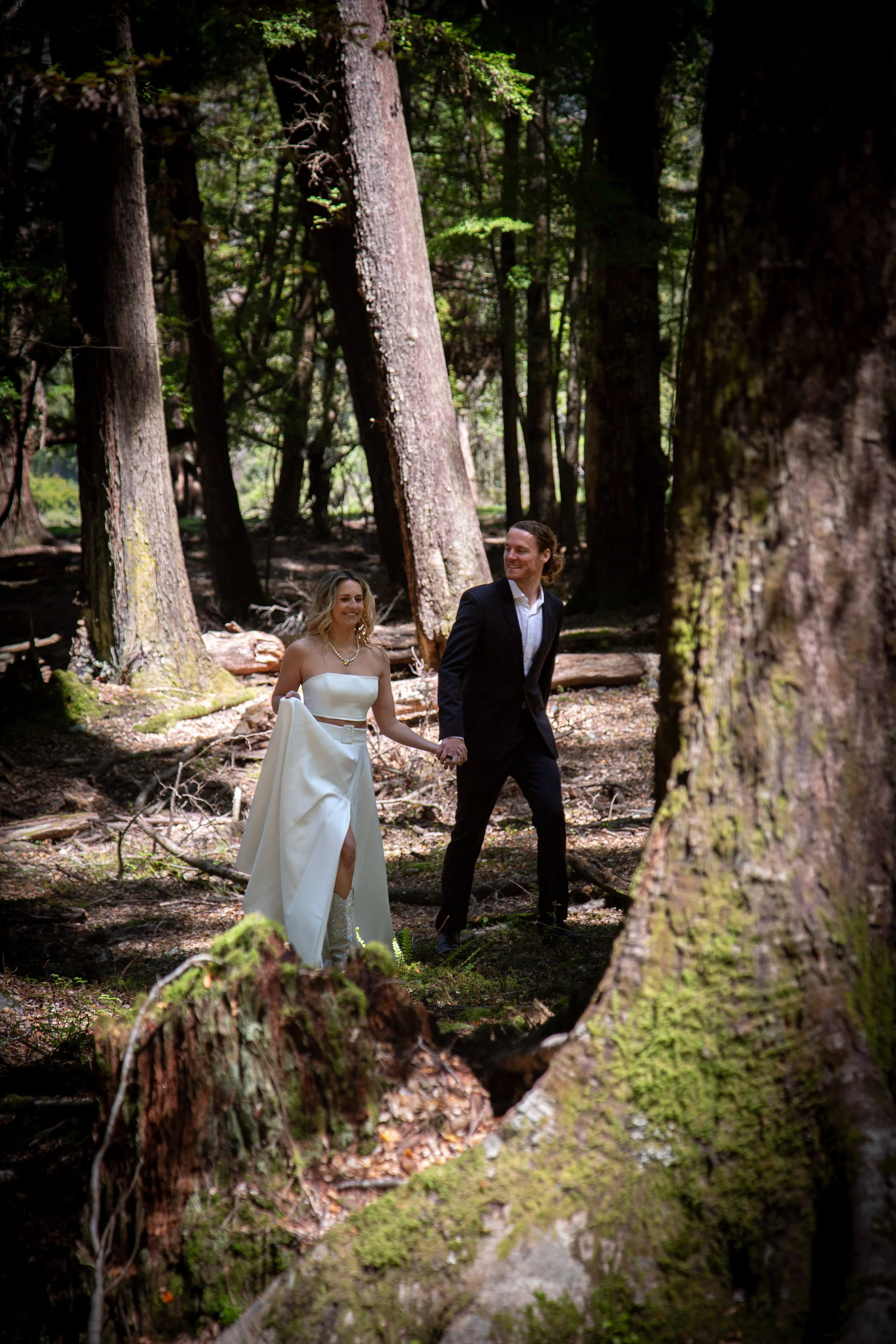 A bride in a white wedding dress and a groom in a black suit walk hand in hand through a forested area.