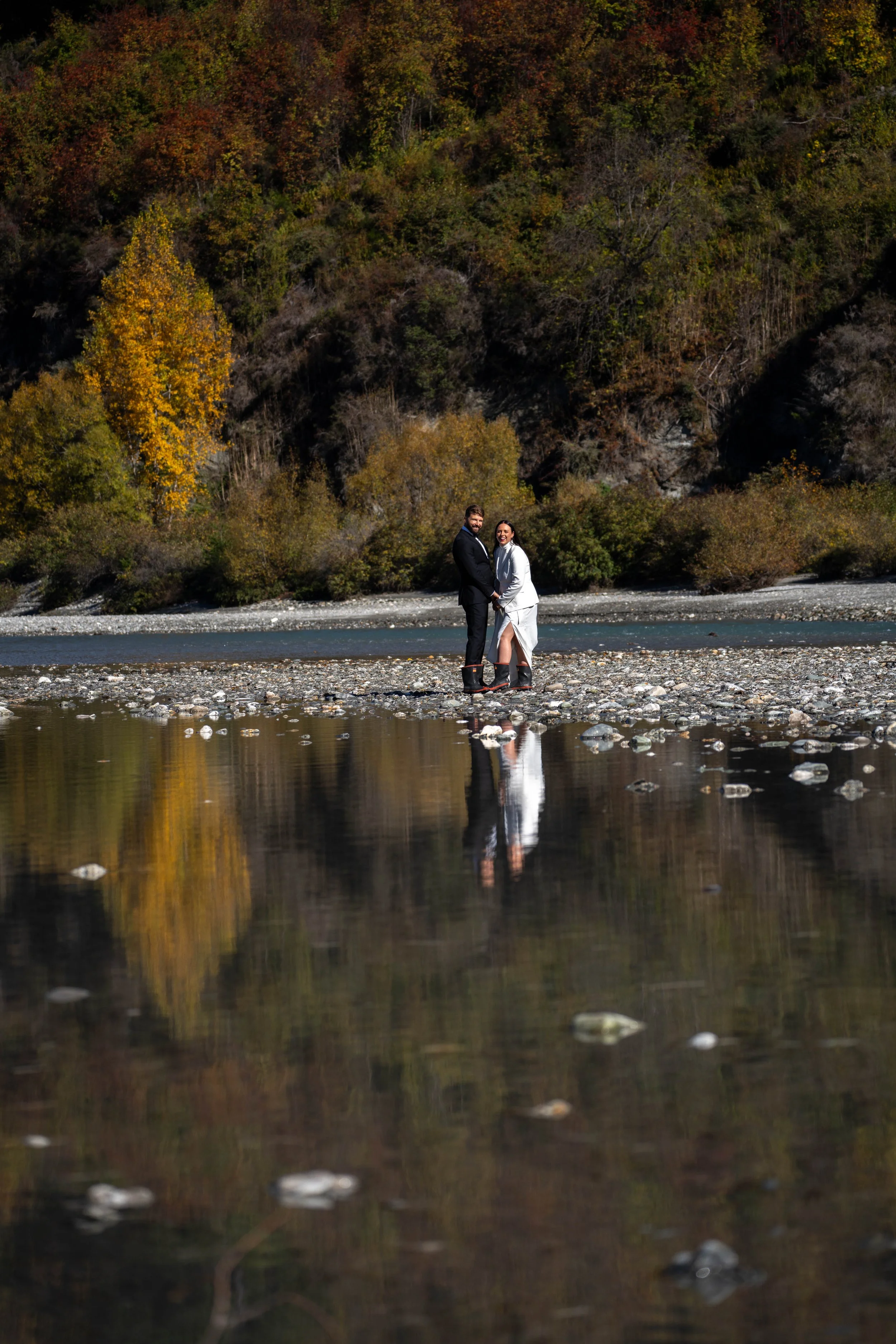 A couple dressed in wedding attire standing on a rocky riverbank with autumn-colored trees and hills in the background, their reflection visible in the water.