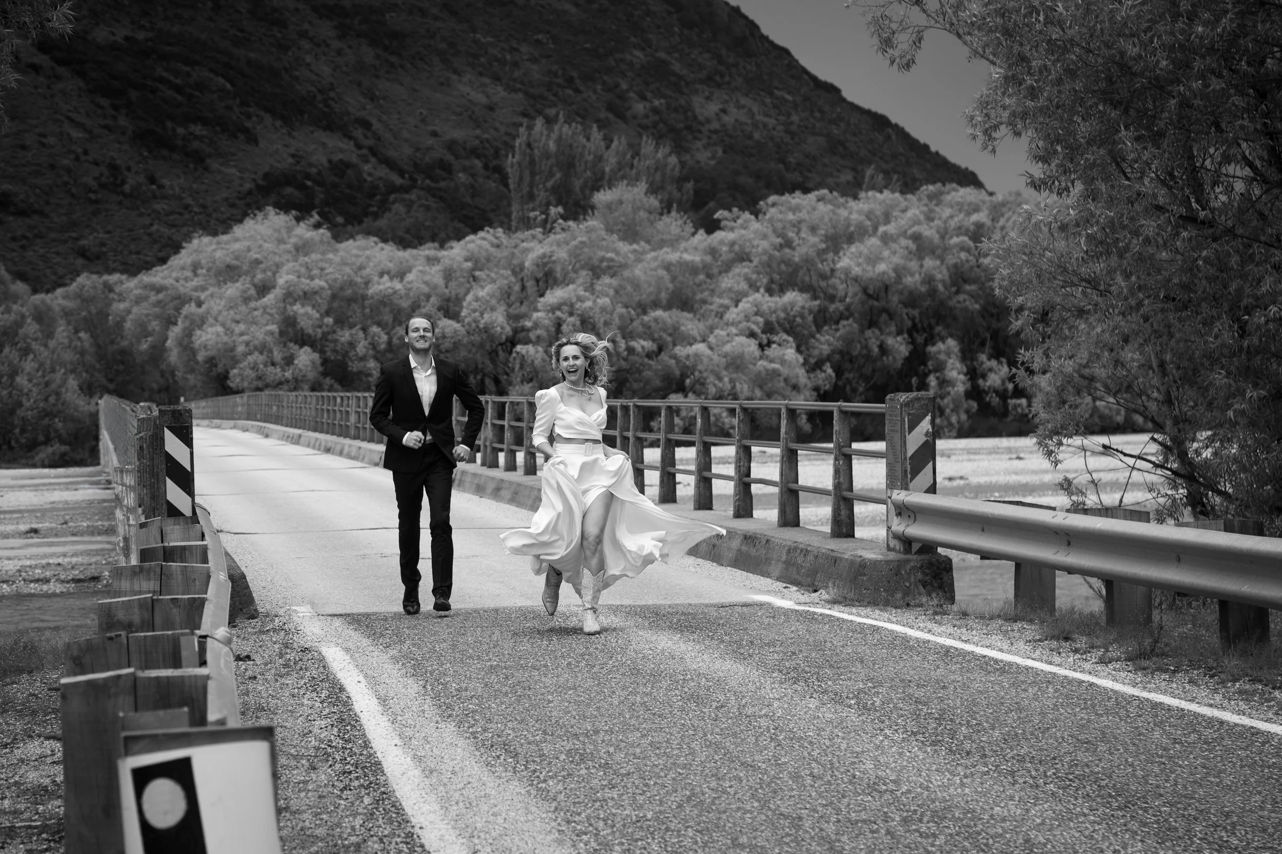 A black-and-white photo of a man and woman running on a bridge in a rural area, with trees and mountains in the background.