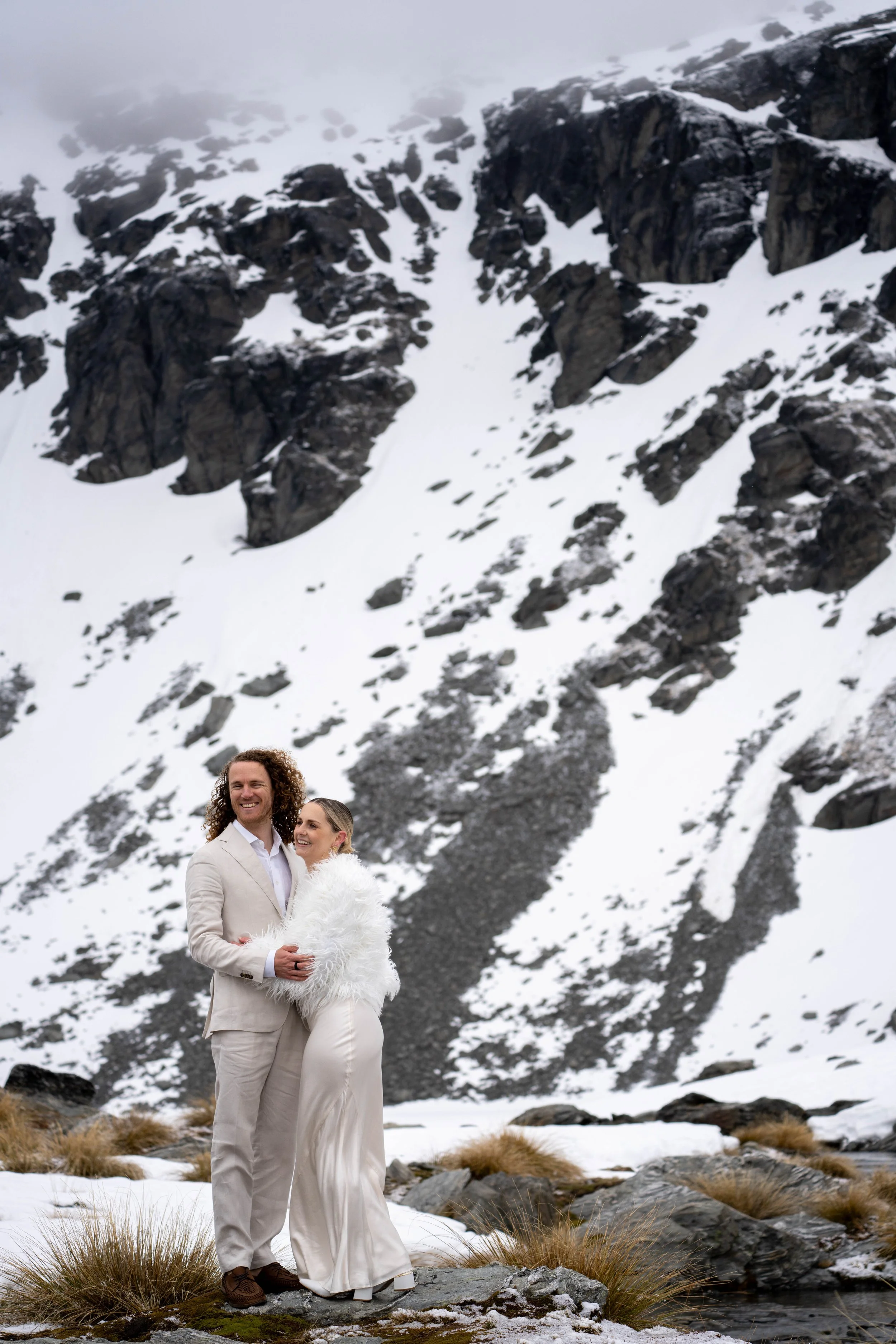 A couple dressed in white standing in a snowy mountain landscape.