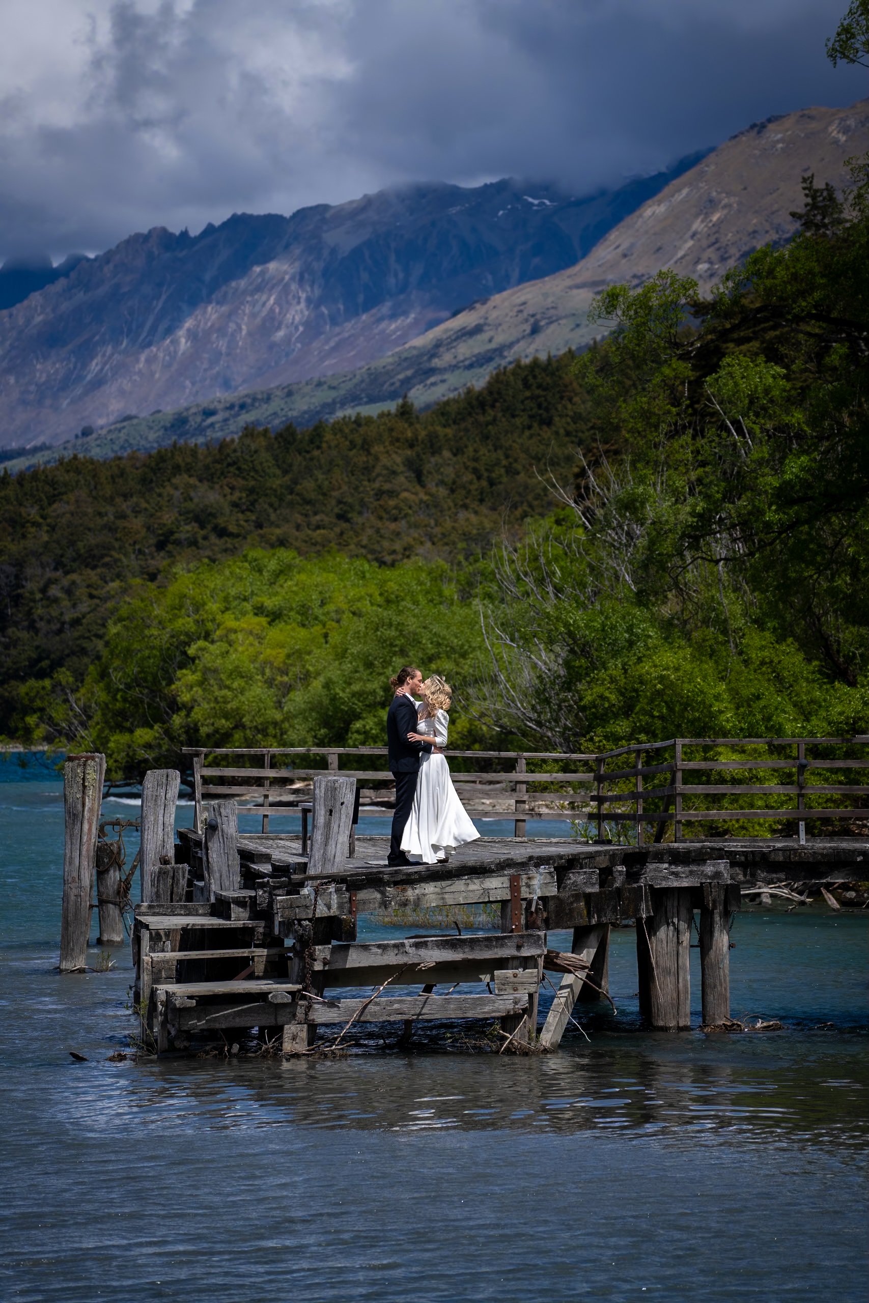 A bride and groom standing on a weathered wooden dock by a lake, embracing against a backdrop of green trees and mountains with cloudy skies.