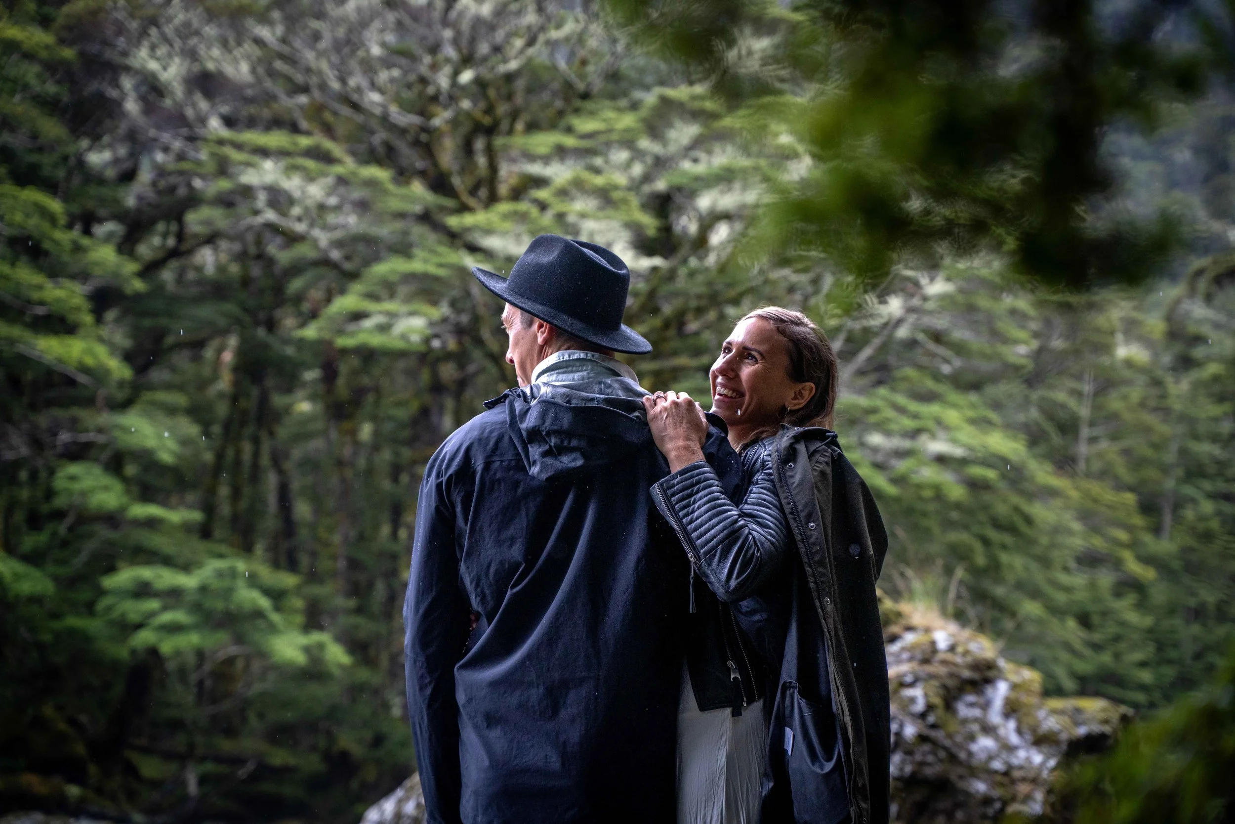 A woman and a man in a forest, the woman is smiling and holding onto the man's shoulder, both wearing outdoor jackets and the man wearing a black hat.
