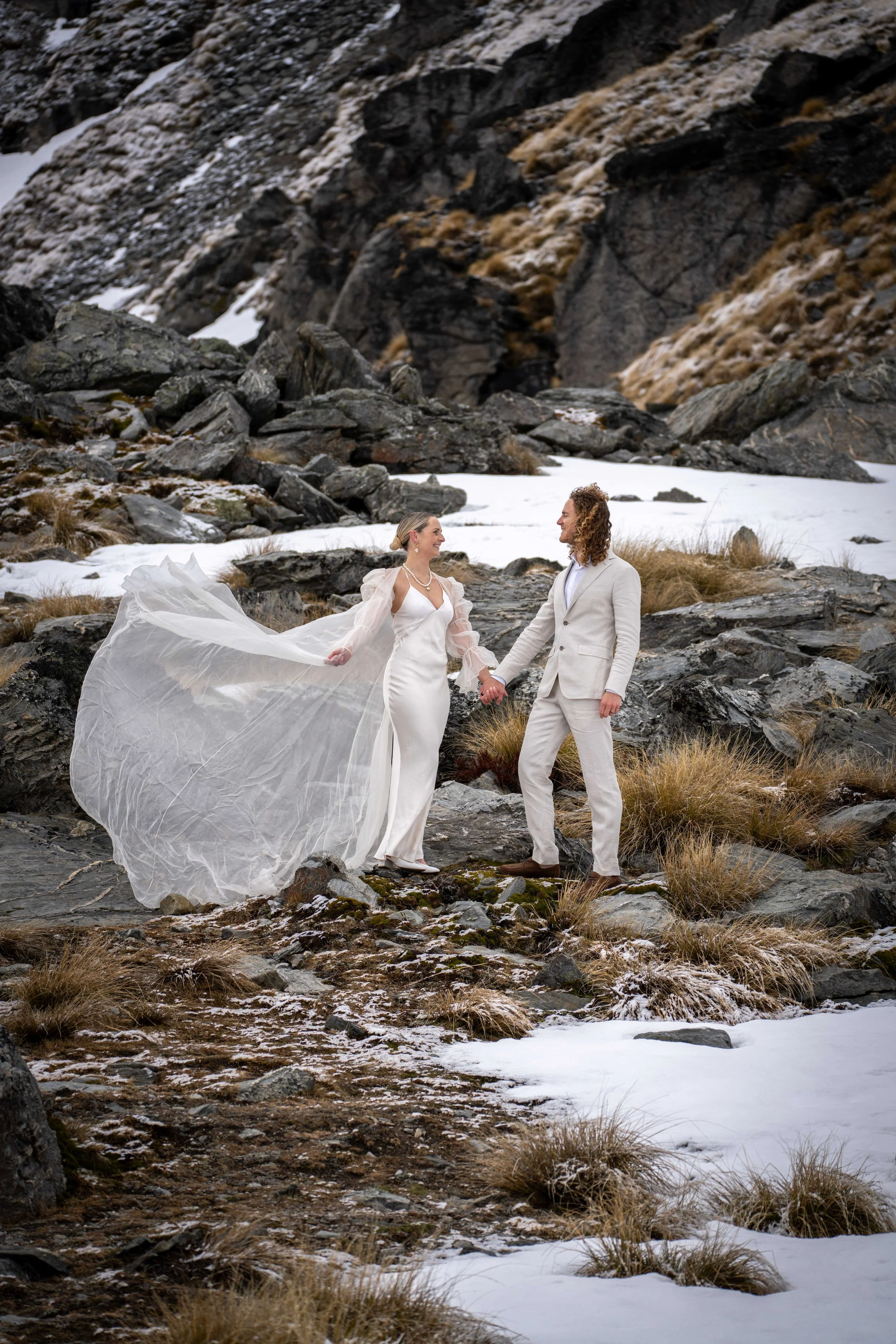 A couple in wedding attire holding hands on rocky terrain with patches of snow and dry grass, mountains in the background.