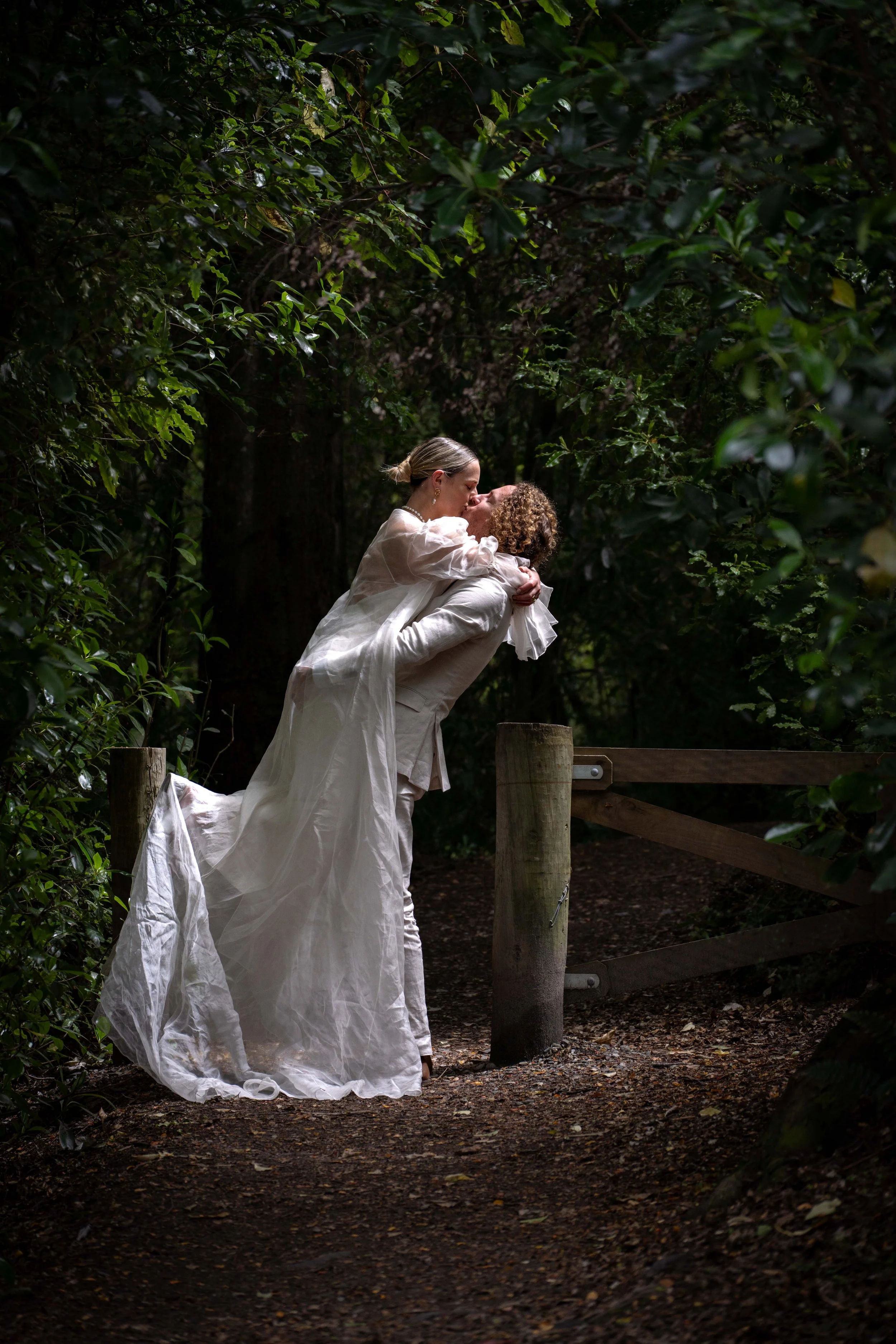 A couple dressed in wedding attire sharing a kiss in a forested area near a wooden fence.