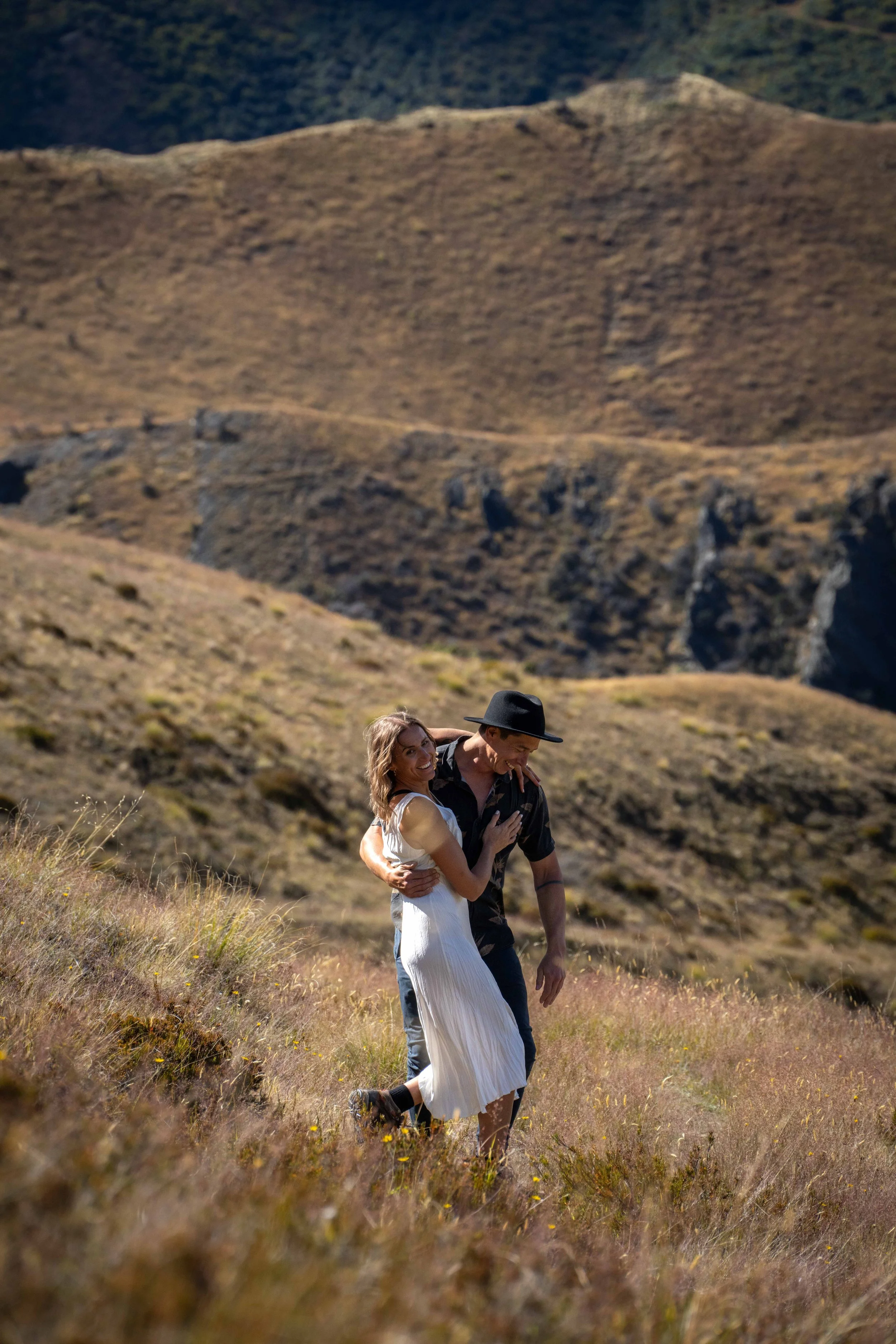 A young couple smiling and embracing while walking through a grassy field in a mountainous landscape.