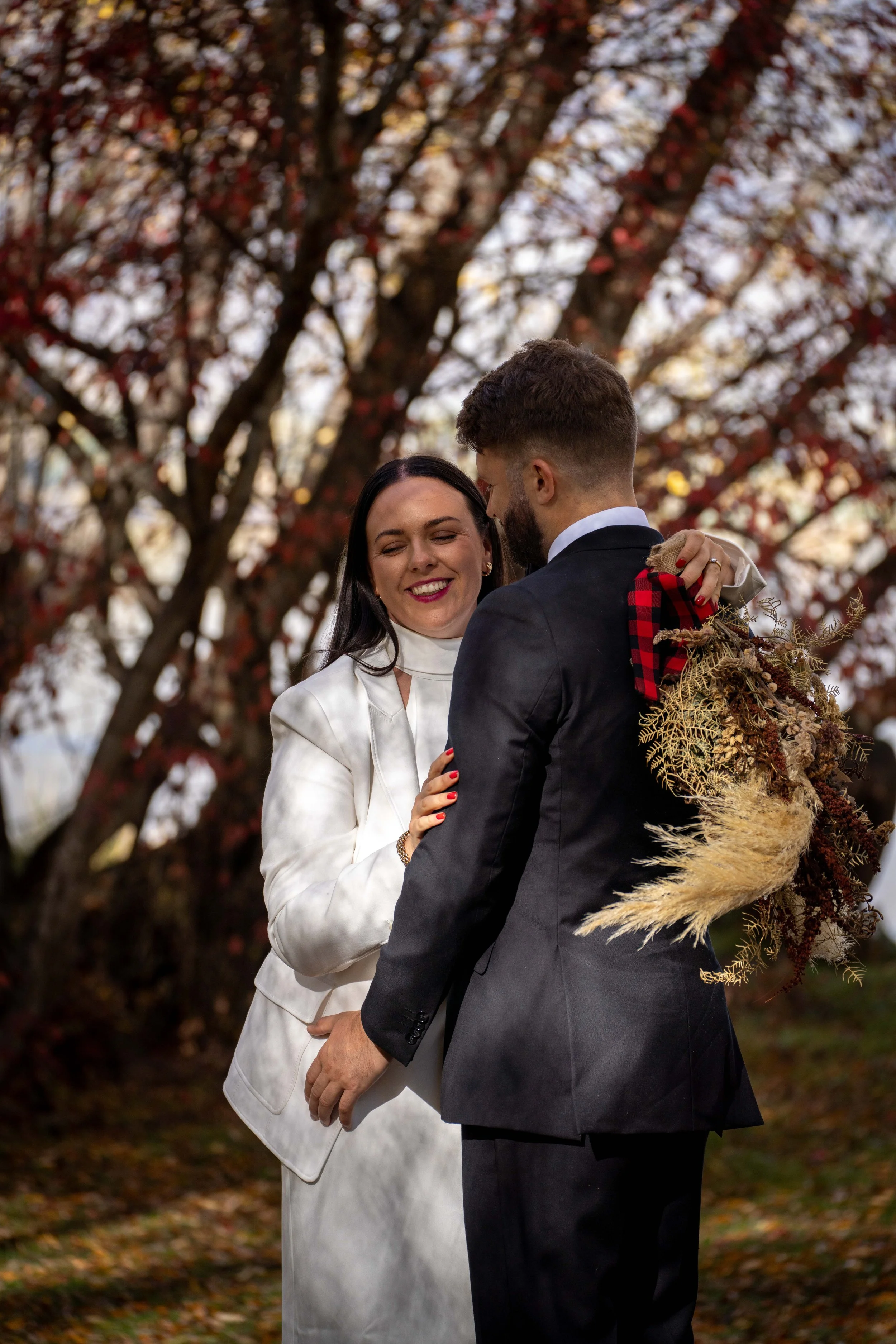 A woman in a white dress and a man in a black suit are standing close together outdoors during autumn, with fallen leaves on the ground and a tree with red and orange leaves in the background. The woman is smiling with her eyes closed, holding a bouq