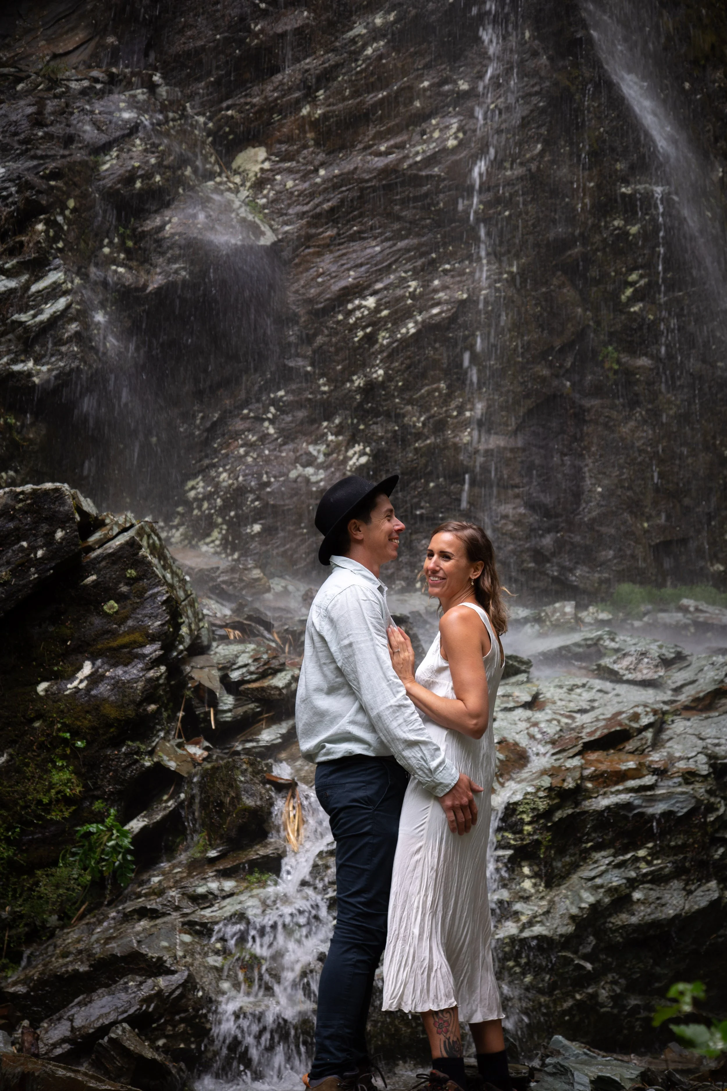 A couple standing under a waterfall, smiling and embracing each other outdoors.