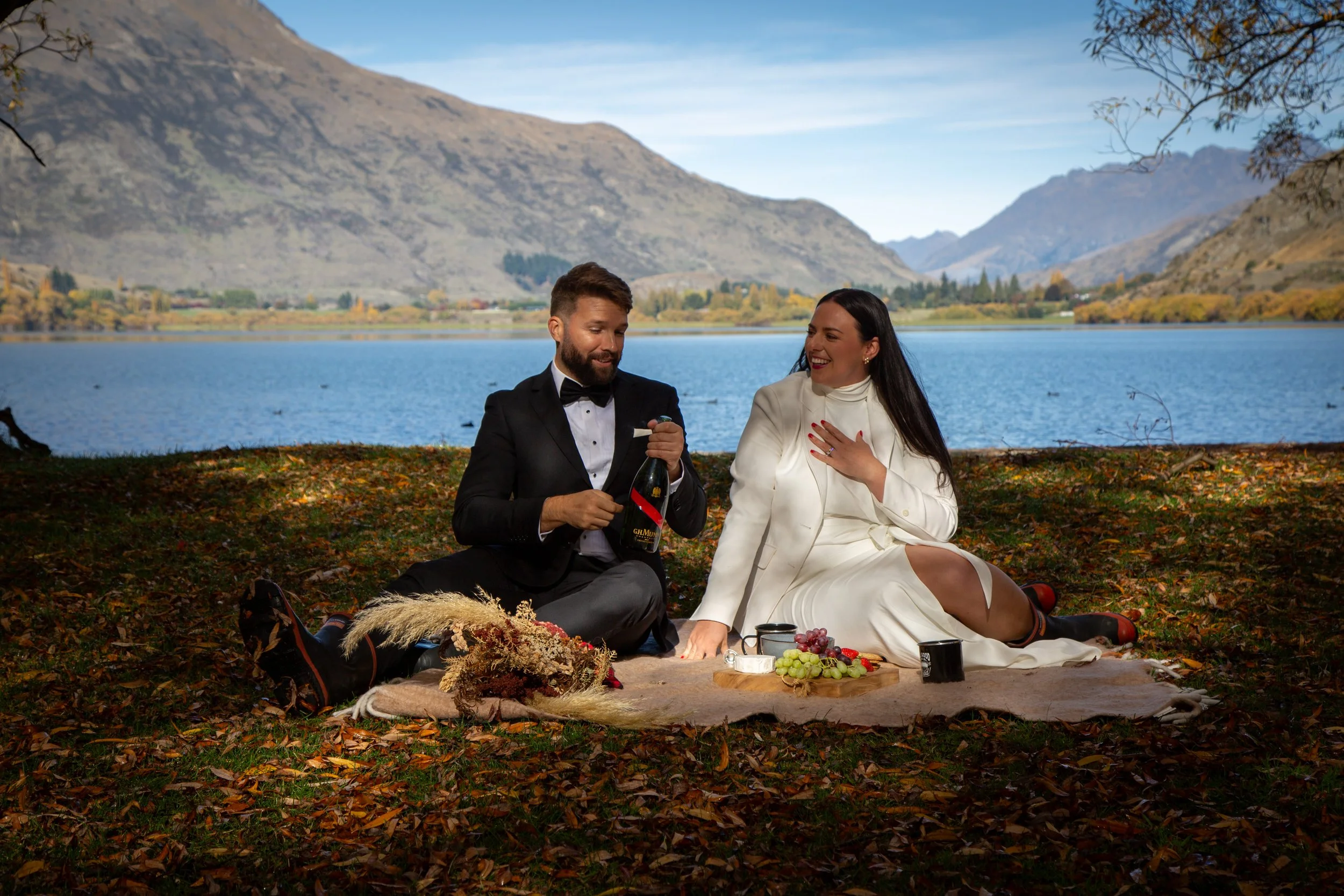 A man and woman dressed in formal attire having a picnic by a lake surrounded by mountains. The man is opening a bottle of champagne, and the woman is sitting on a blanket with a platter of grapes, mugs, and other food.