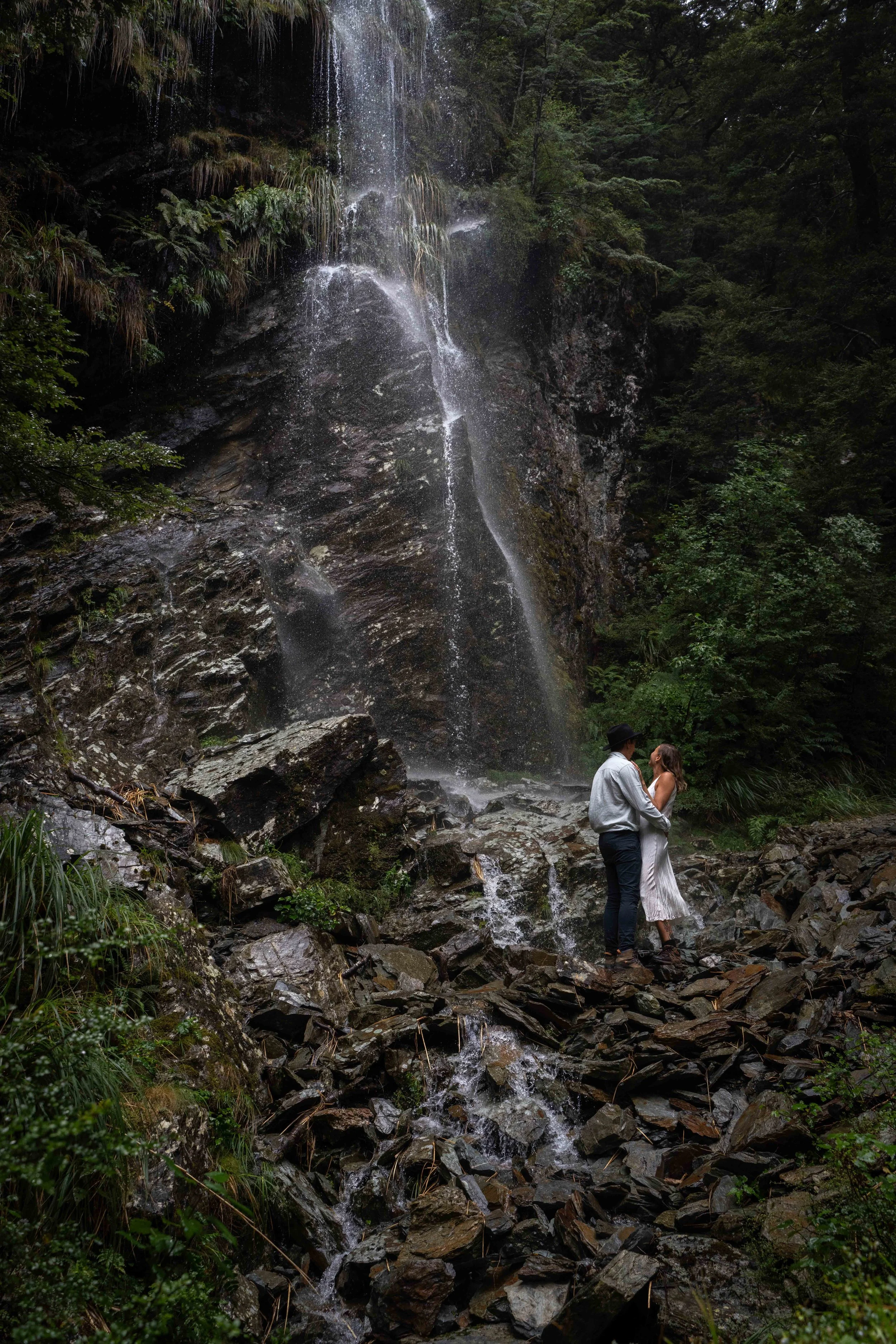 A couple standing on rocks near a waterfall in a forest.