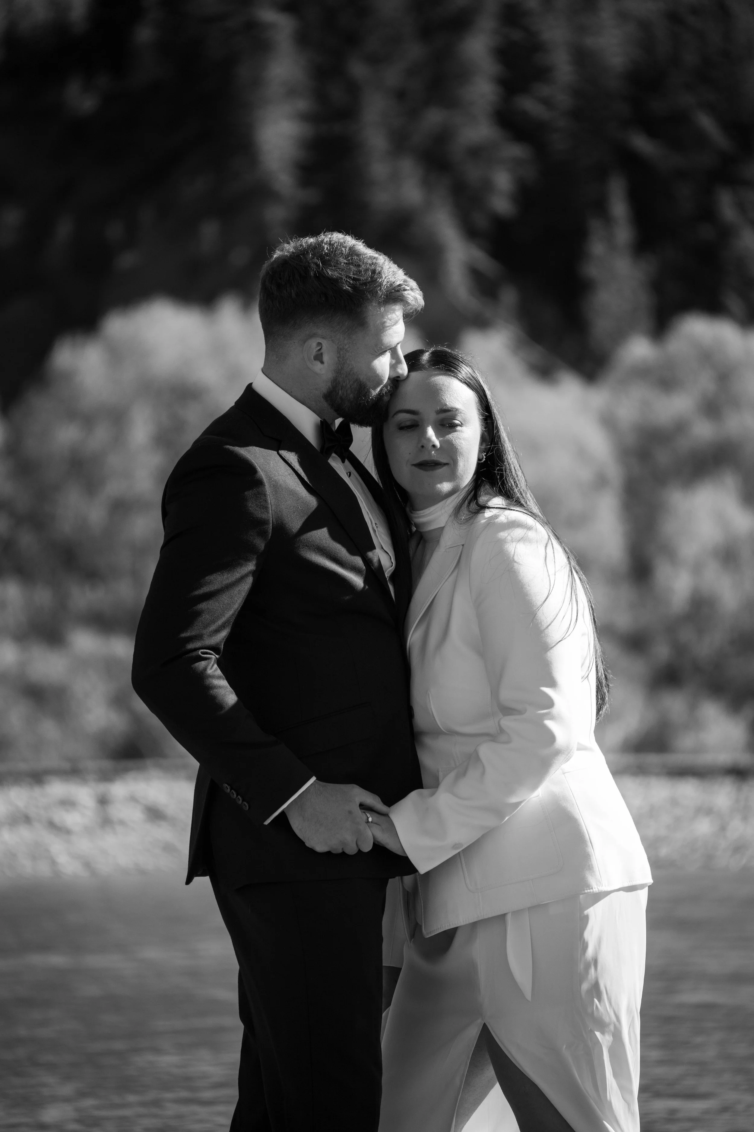 A black and white photo of a couple in formal attire sharing an intimate moment outdoors by a body of water, with trees in the background.