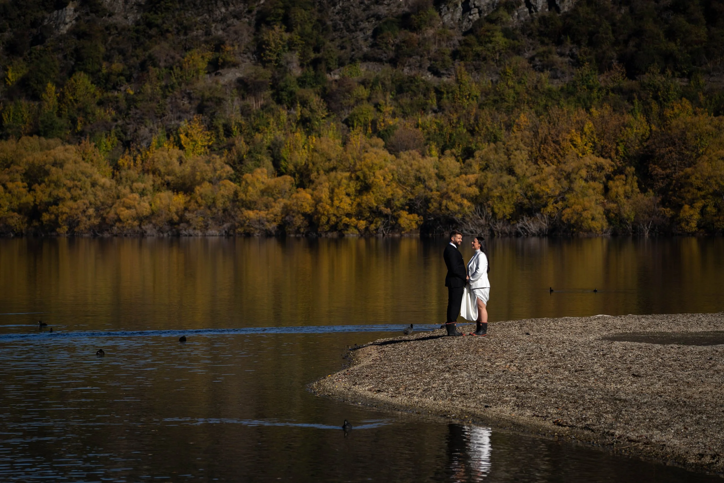 A couple in wedding attire holding hands on a pebble beach at the edge of a calm lake, with a forested hillside showing fall foliage in the background.